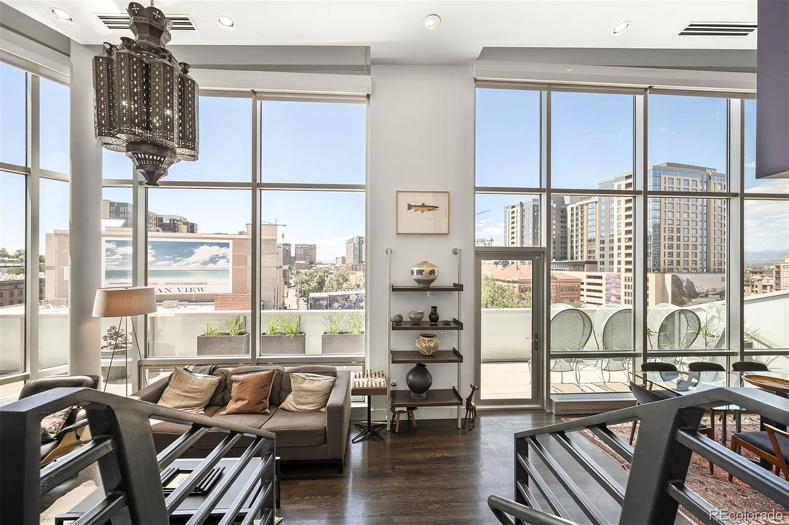 This is an interior shot of a modern living room featuring floor-to-ceiling windows that offer a stunning city view. The room is furnished with a comfortable sofa, decorative pillows, and a unique chandelier. A staircase with metal railings adds an architectural element to the space, and the dark hardwood floors provide a rich contrast to the light walls.