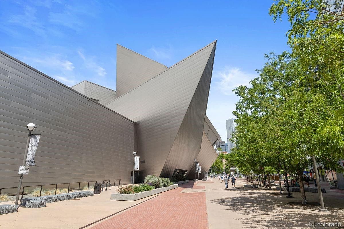 This image showcases the side of a modern architectural building with a unique, angular design and metallic cladding. The building is situated along a pedestrian walkway lined with trees, creating a visually appealing urban landscape. The clear blue sky enhances the overall impression of a well-maintained and architecturally significant property.