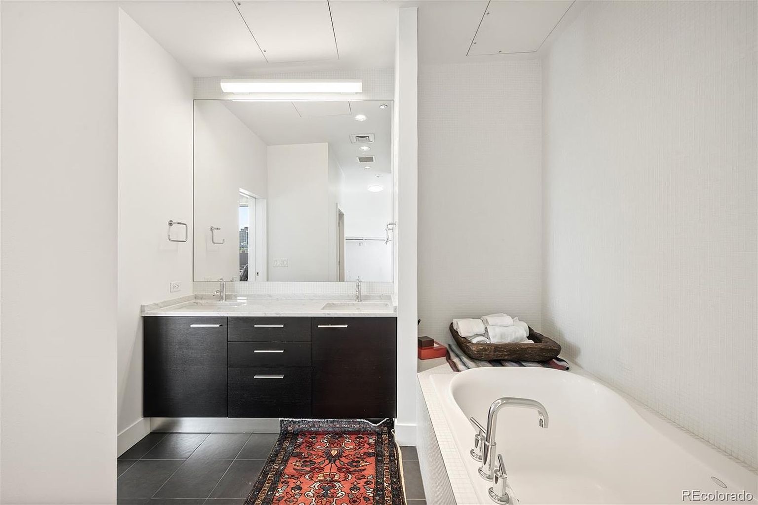This is a well-lit primary bathroom featuring a double vanity with dark wood cabinets and a marble countertop. A large mirror spans the length of the vanity, reflecting the bright lighting. A modern bathtub with white tiling and a chrome faucet is positioned to the right, complemented by neatly arranged white towels, creating a spa-like atmosphere.