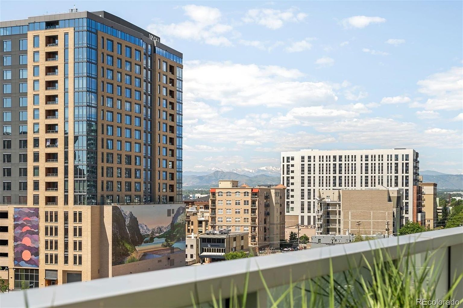 This image showcases a cityscape view from a patio or balcony, featuring modern high-rise buildings with glass and brick facades. In the background, there are snow-capped mountains, adding a scenic element to the urban landscape. The view suggests a desirable location with a blend of city and natural beauty.