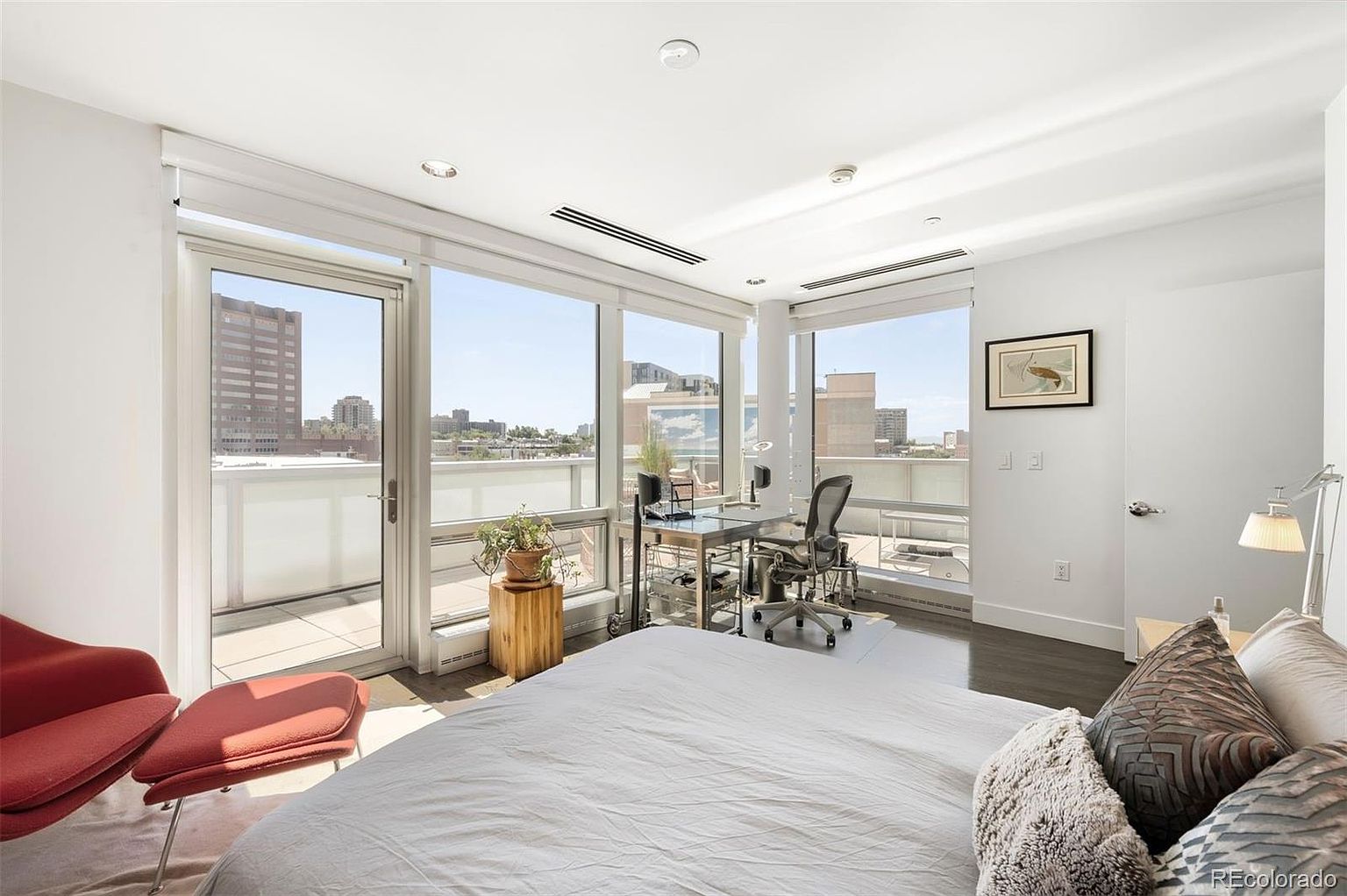 This is a bright and airy primary bedroom featuring floor-to-ceiling windows that offer city views and access to a balcony. The room includes a modern desk area, a red lounge chair, and a bed with decorative pillows. The overall aesthetic is contemporary and minimalist, emphasizing natural light and urban living.