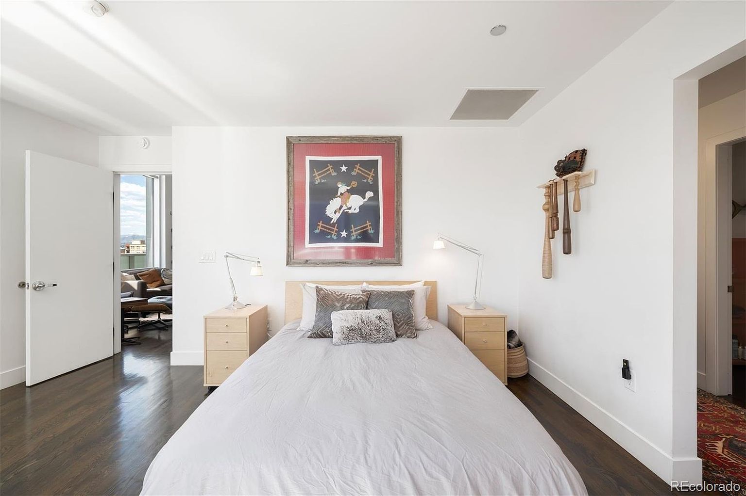 This is a well-lit primary bedroom featuring a queen-sized bed with neutral bedding and decorative pillows. Two matching nightstands with lamps flank the bed, and a framed artwork hangs above the headboard. The room has dark hardwood floors and white walls, creating a clean and modern aesthetic.