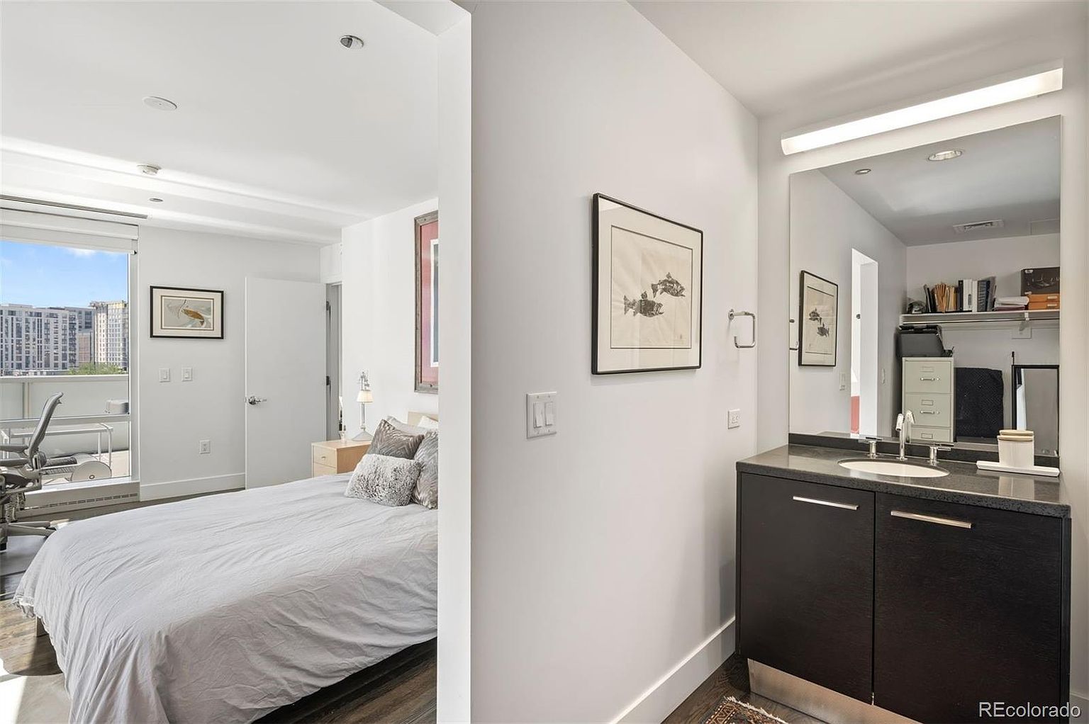 This image showcases a primary bedroom connected to a bathroom. The bedroom features a large bed with neutral bedding, a window with a city view, and a desk area. The bathroom has a modern vanity with a dark cabinet and a large mirror, reflecting a closet area. The overall impression is clean, contemporary, and well-lit.