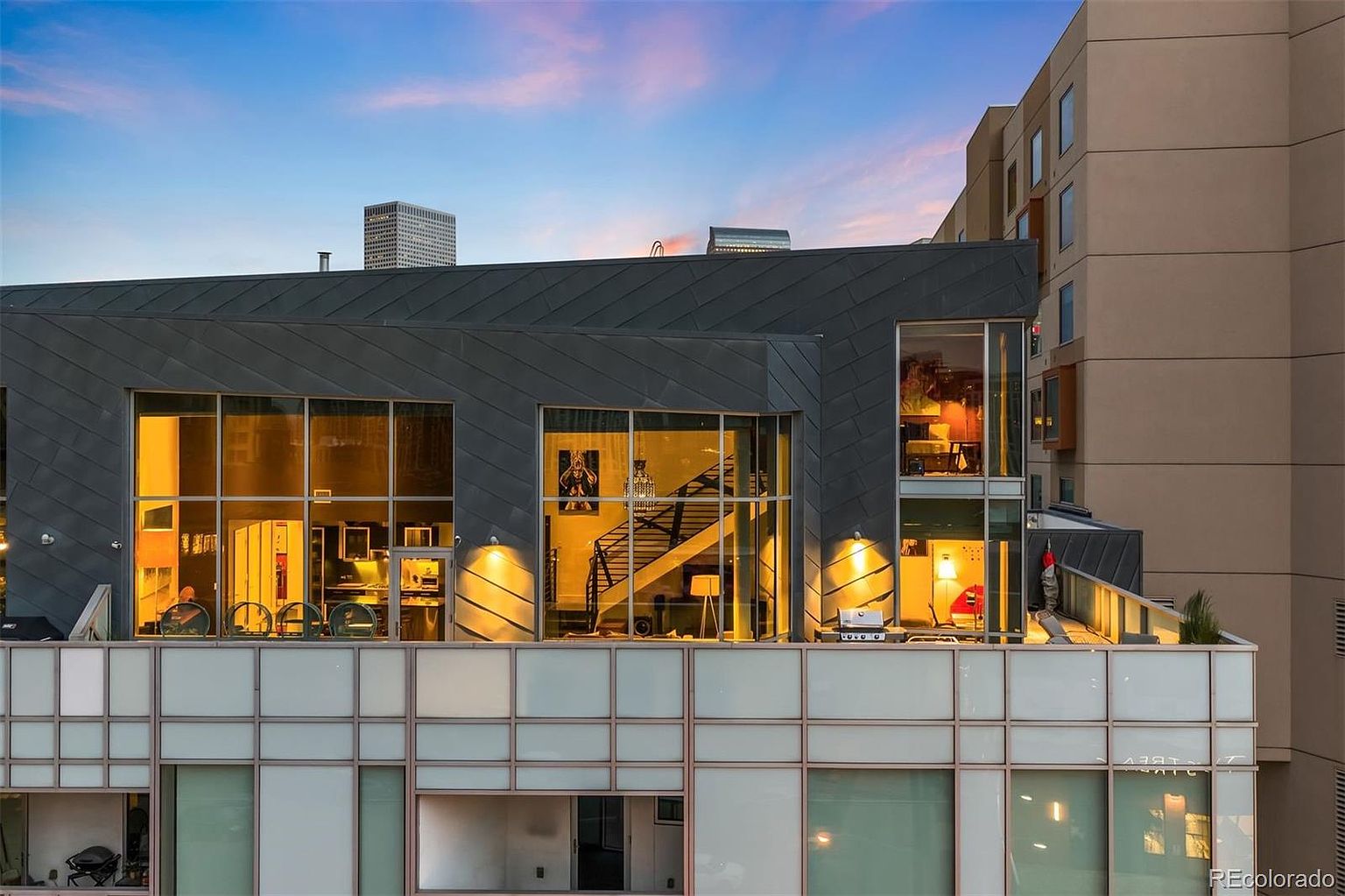 This image showcases a modern building exterior with a prominent patio/deck/balcony area. The structure features large windows offering views into the interior spaces, including a staircase and living areas. The balcony is equipped with outdoor furniture and a grill, suggesting an inviting outdoor living space. The building's architecture is contemporary, with a combination of gray and white panels, and the overall impression is one of urban sophistication.