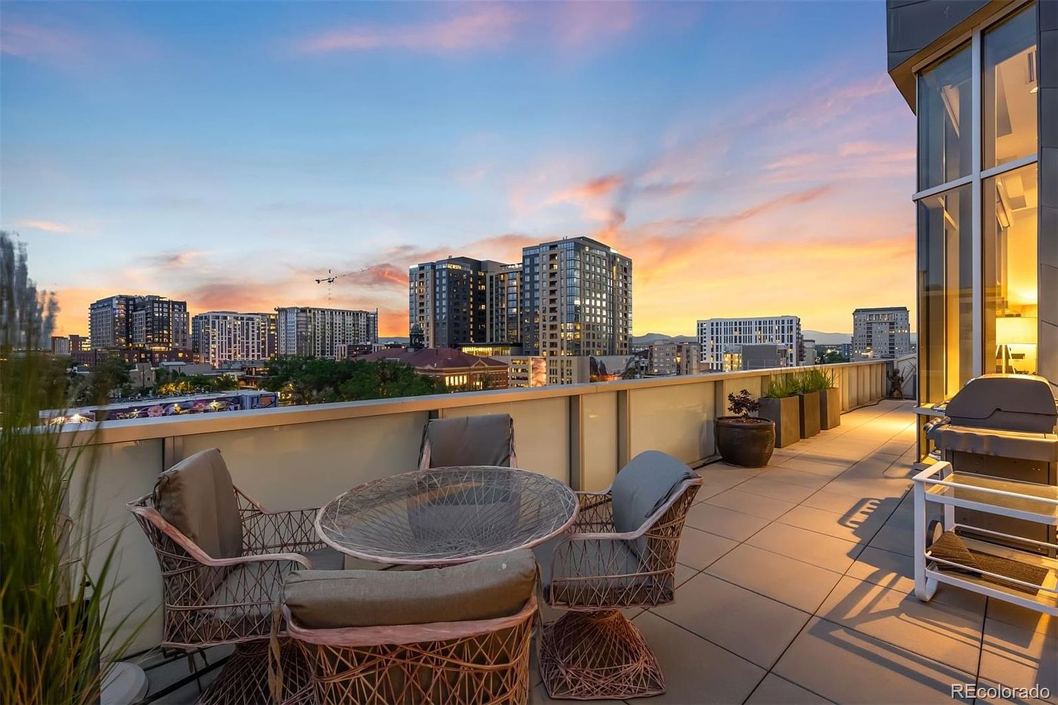 This image showcases a luxurious rooftop patio or balcony with stunning city views at sunset. The space is furnished with stylish wicker seating around a glass-topped table, creating an inviting outdoor living area. A modern grill and sleek planters add to the upscale ambiance, making it an ideal spot for relaxation and entertaining.