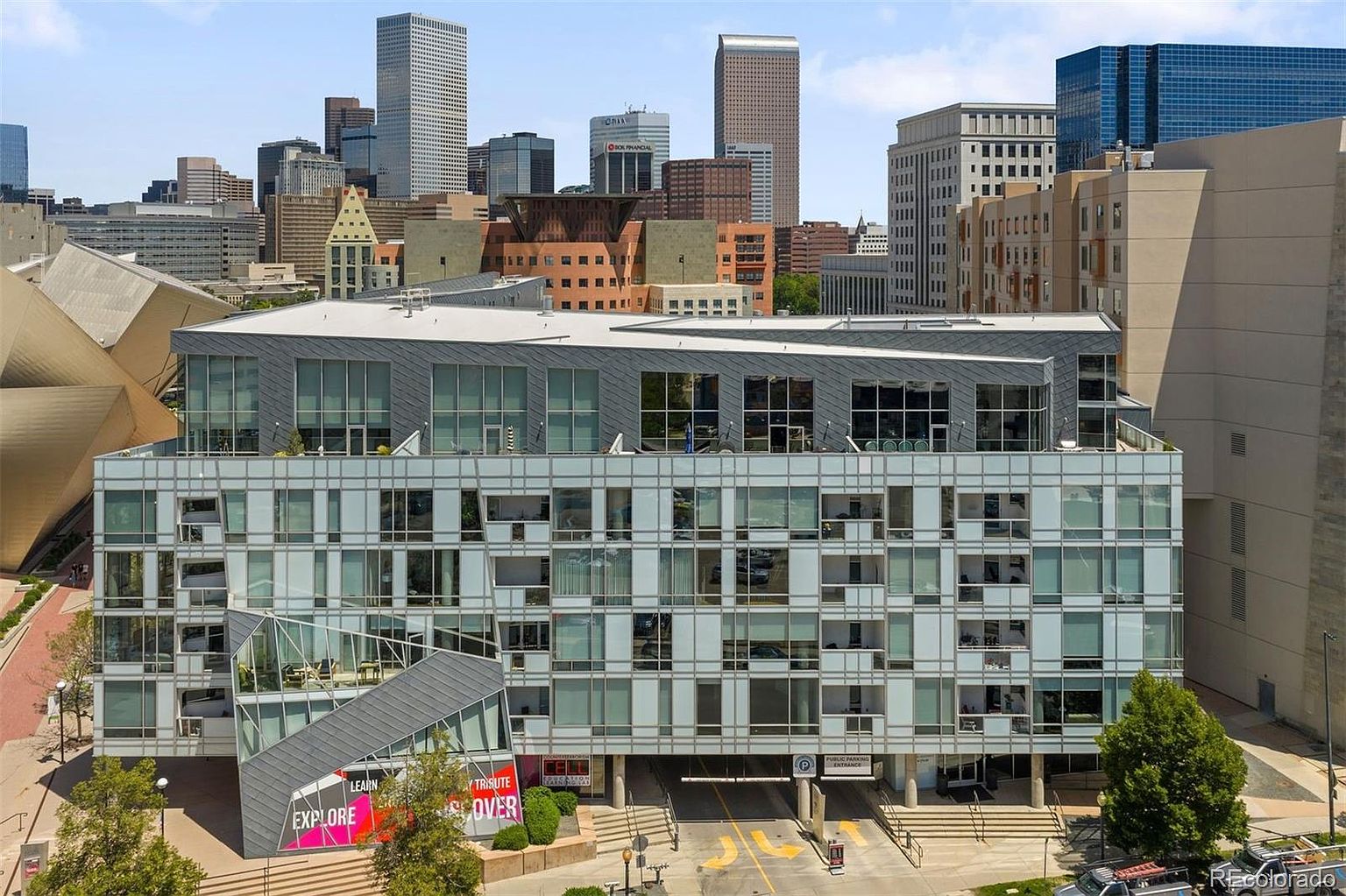 This is a front view of a modern, multi-story building with a grid-like facade of windows and metal accents. The building features a unique architectural design with a prominent entrance and signage. The surrounding cityscape includes other high-rise buildings, creating an urban environment.