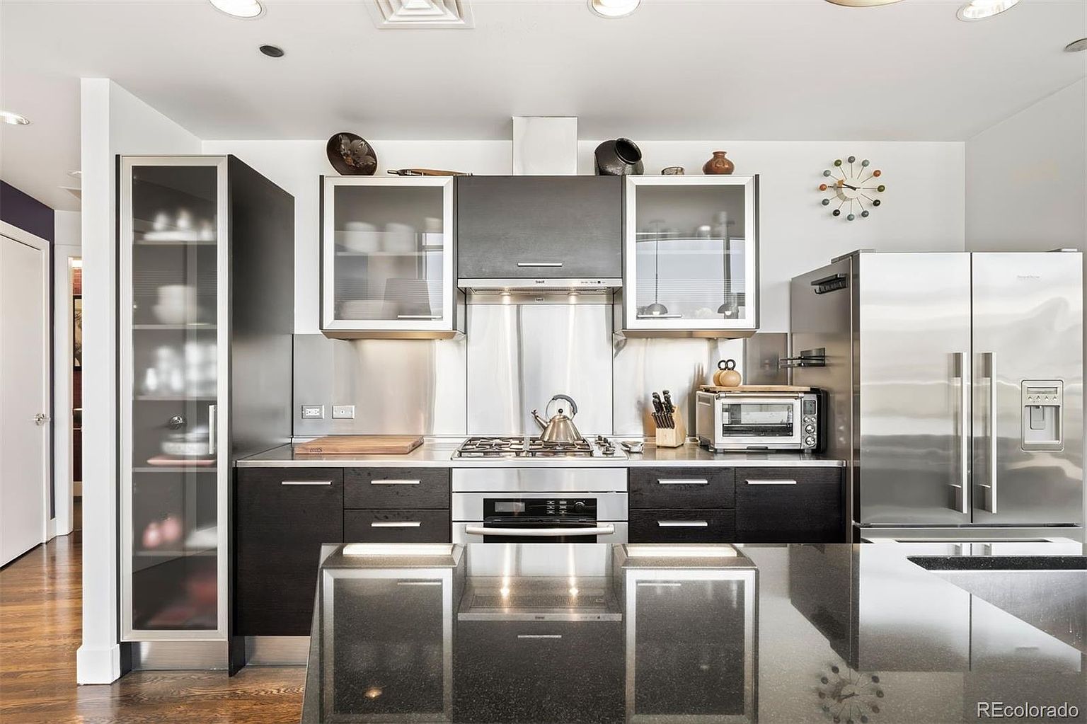This is a modern kitchen featuring dark wood cabinetry, stainless steel appliances, and a black countertop island. The kitchen is well-lit with recessed lighting and under-cabinet lighting, highlighting the stainless steel backsplash and the glass-fronted upper cabinets. The overall impression is sleek and contemporary.