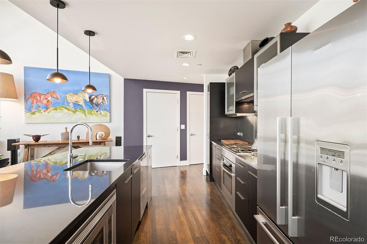 This is a modern kitchen with dark cabinetry, stainless steel appliances, and hardwood floors. A large island with a dark countertop dominates the center of the room, reflecting the pendant lights above. The kitchen also features a stainless steel refrigerator and a gas cooktop.