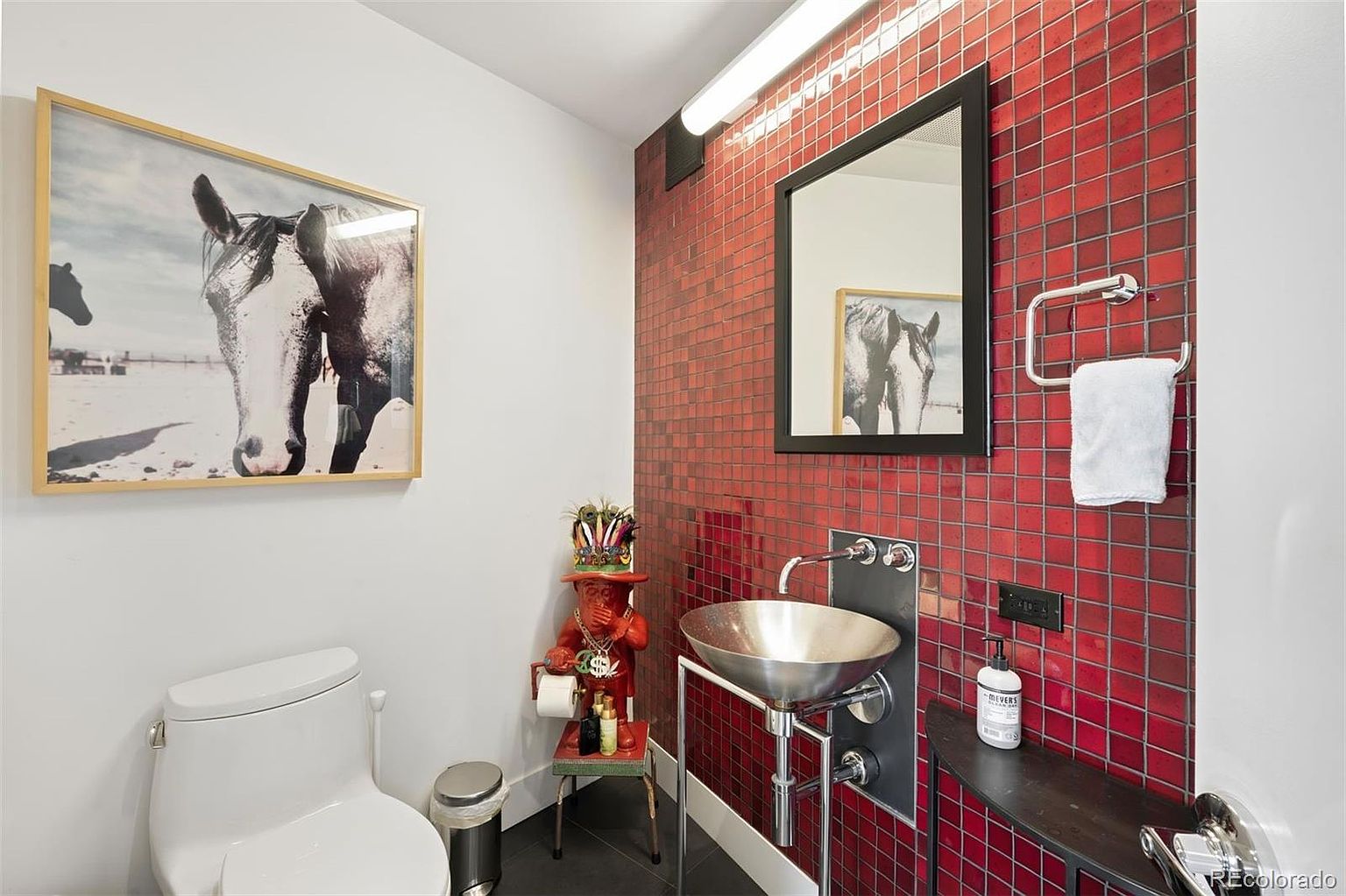 This is a stylish guest bathroom featuring a unique design. The walls are adorned with red mosaic tiles, complemented by a modern, vessel sink and a black-framed mirror. A framed horse print adds a touch of character, while a quirky statue provides a playful element to the space.