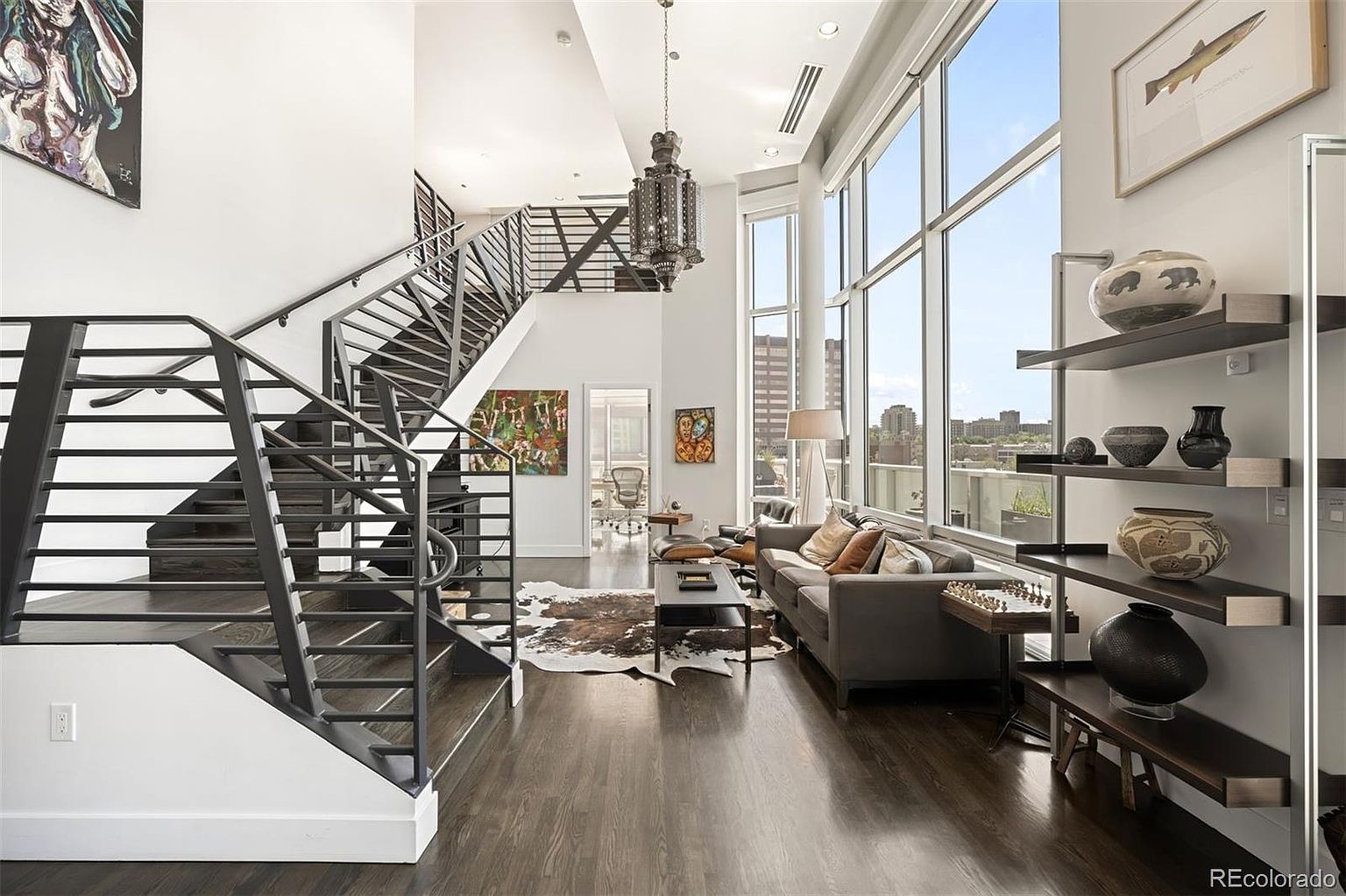 This is a spacious and modern living room featuring a striking staircase with metal railings, hardwood floors, and floor-to-ceiling windows offering city views. The room is furnished with a gray sofa, a coffee table, and unique shelving displaying decorative items. The overall impression is one of contemporary elegance and urban sophistication.