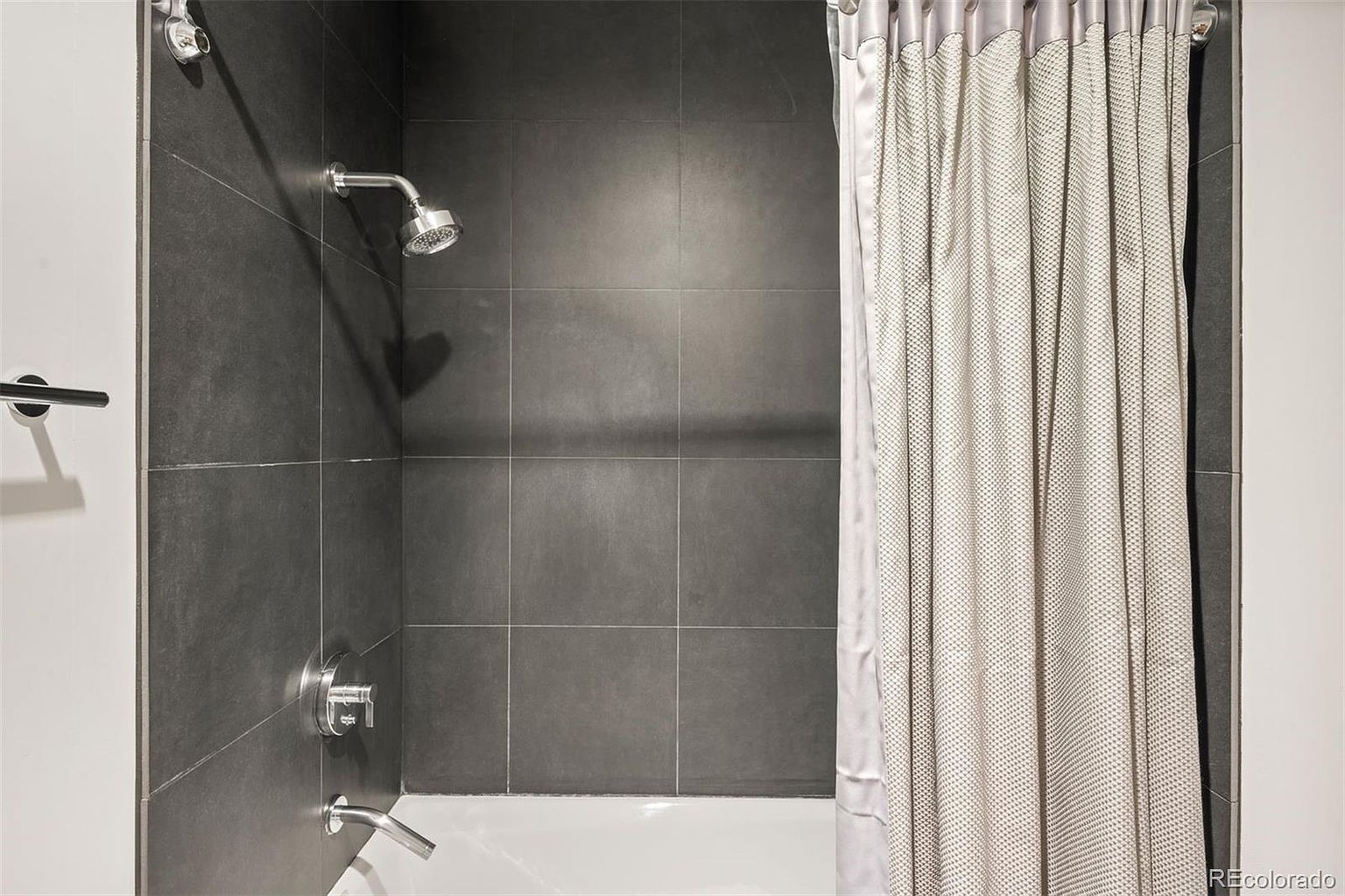 This is a bathroom featuring a shower with dark gray tiled walls and a white shower curtain. The shower has chrome fixtures, including a shower head, faucet, and handle. The overall impression is modern and clean, with a focus on functionality.