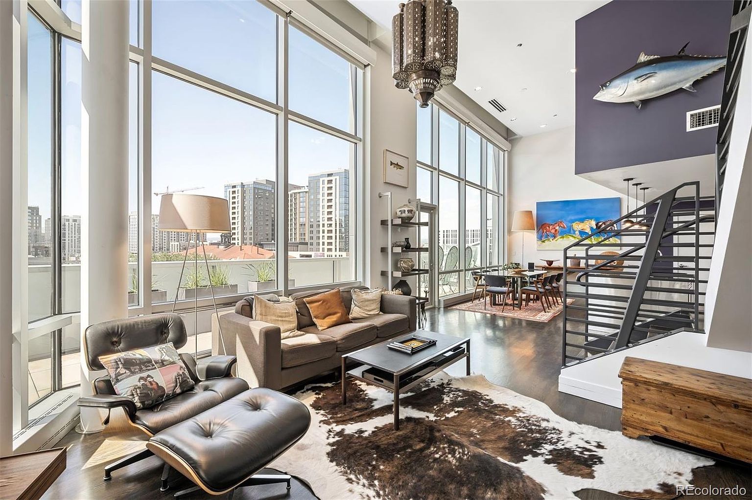 This is a spacious and modern living room with floor-to-ceiling windows offering city views. The room features a comfortable gray sofa, a black leather chair with ottoman, and a unique cowhide rug. A staircase leads to an upper level, and a large decorative fish hangs on the wall, adding a touch of whimsy.