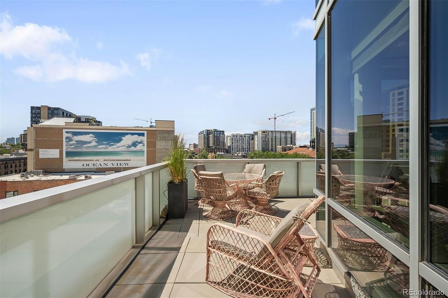 This image showcases a stylish balcony or patio area with modern outdoor furniture, including wicker chairs and a glass-topped table. The space offers a cityscape view, with buildings and construction cranes visible in the background. The balcony features a sleek railing and tiled flooring, creating a sophisticated outdoor living space.