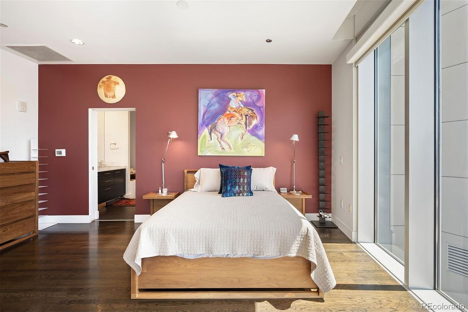 This is a primary bedroom featuring a wooden bed with a white quilt and a blue decorative pillow. The room has a warm, inviting feel with a red accent wall adorned with artwork and bedside lamps. Large windows provide natural light, and the hardwood floors add to the room's elegance.