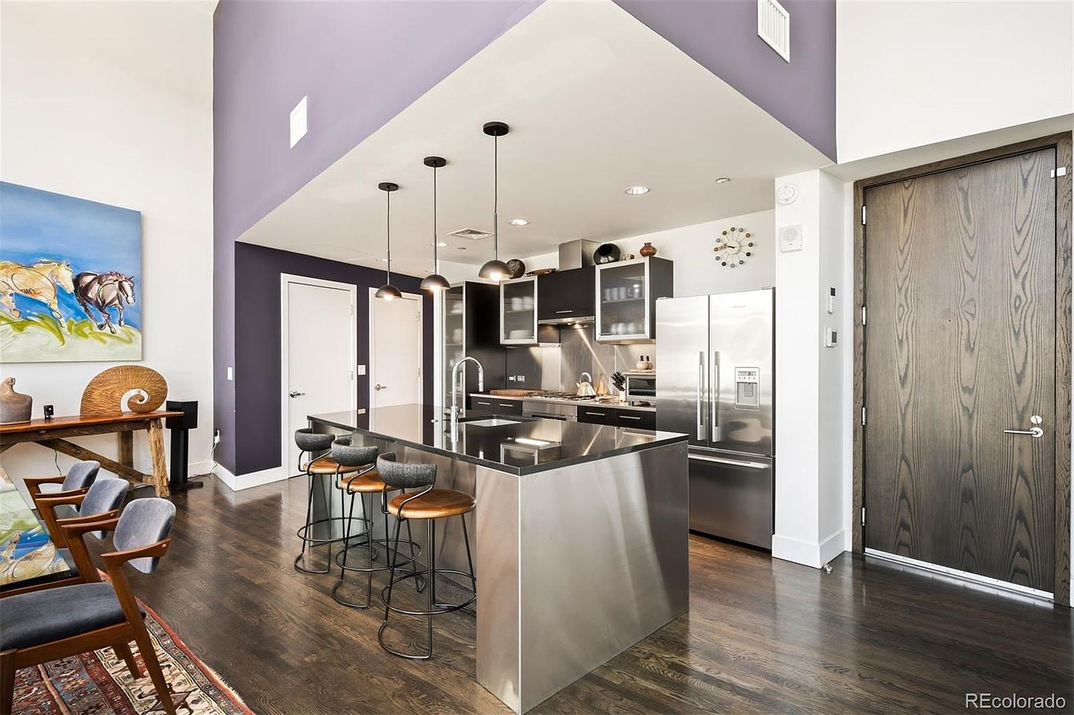 This interior shot showcases a modern kitchen with dark wood floors, a stainless steel island with seating, and sleek black cabinetry. Stainless steel appliances, including a refrigerator, complement the contemporary design. The open layout connects to a living area, highlighted by a large painting, creating a stylish and inviting space.