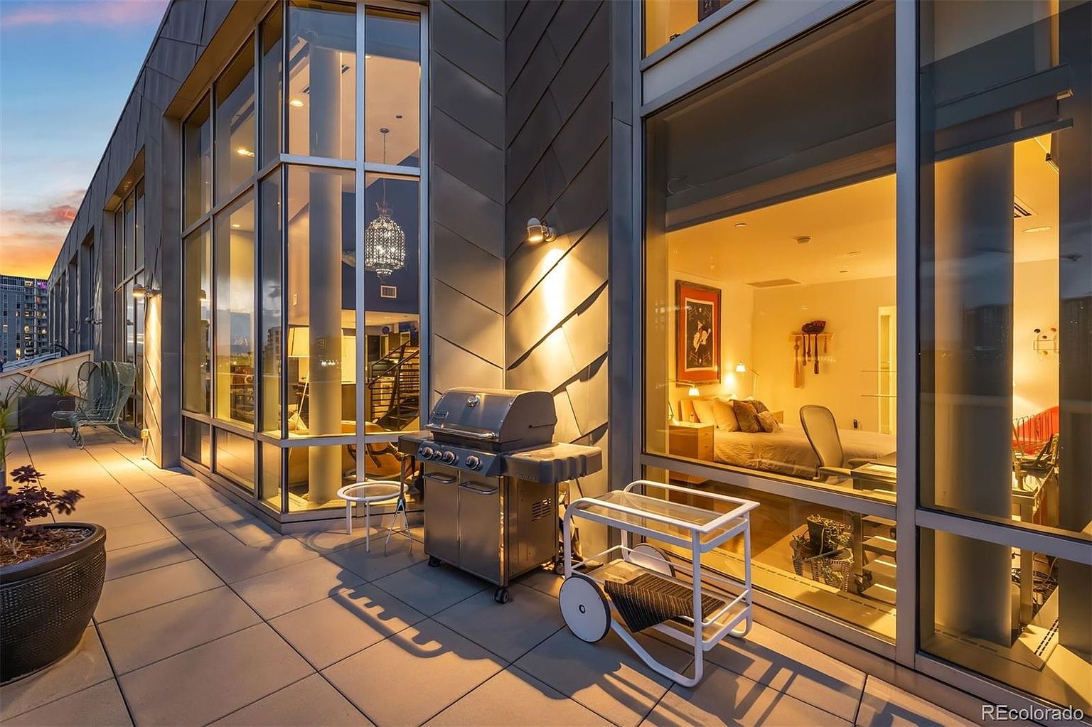 This image showcases a luxurious patio or balcony area, featuring a modern grill, a serving cart, and stylish outdoor seating. The space is defined by large windows offering views into the interior living spaces, including a bedroom and a living area with a staircase. The overall impression is one of upscale urban living with a focus on outdoor entertaining.