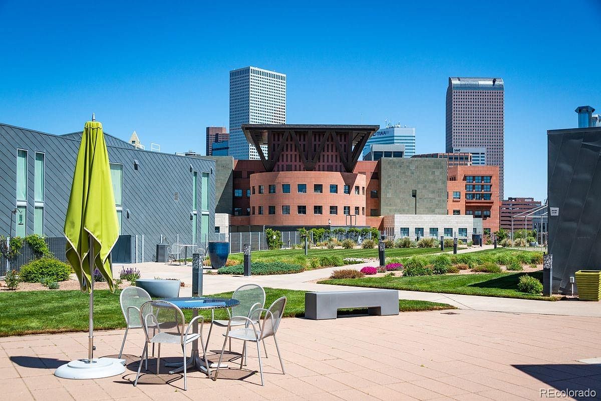 This image showcases a modern patio or deck area with a table and chairs, an umbrella, and a bench, set against a backdrop of contemporary buildings and a clear blue sky. The well-maintained lawn and landscaping add to the appeal, creating a pleasant outdoor space. The scene suggests a relaxing and inviting atmosphere, perfect for enjoying the urban landscape.