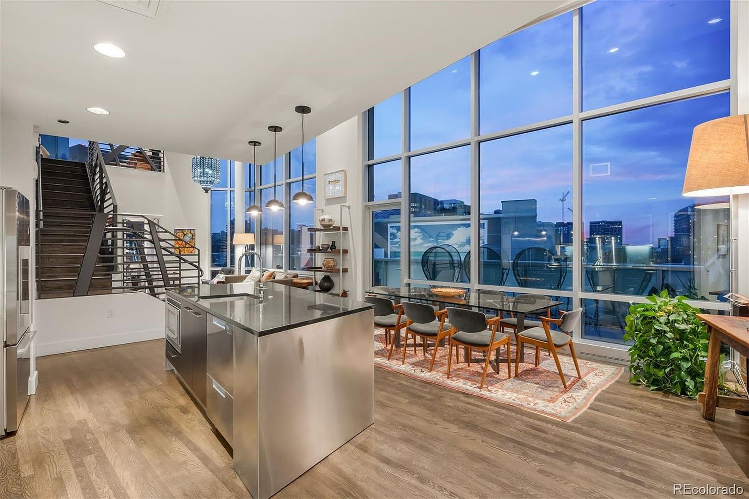 This is a modern kitchen and dining area featuring a stainless steel kitchen island with a black countertop, complemented by stainless steel appliances. The dining area includes a glass-top table with six chairs, set against a backdrop of large windows offering city views. A staircase is visible on the left, adding to the open and contemporary feel of the space.