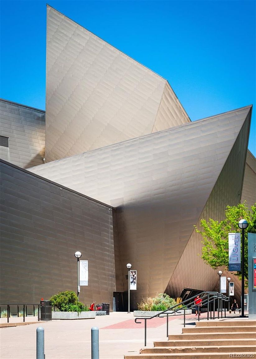 This image showcases the striking front exterior of a modern architectural building, possibly a museum or cultural center. The building features sharp, angular designs with metallic cladding, creating a bold and contemporary aesthetic. The surrounding plaza includes landscaping, street lamps, and pedestrian access, enhancing the building's presence and accessibility.