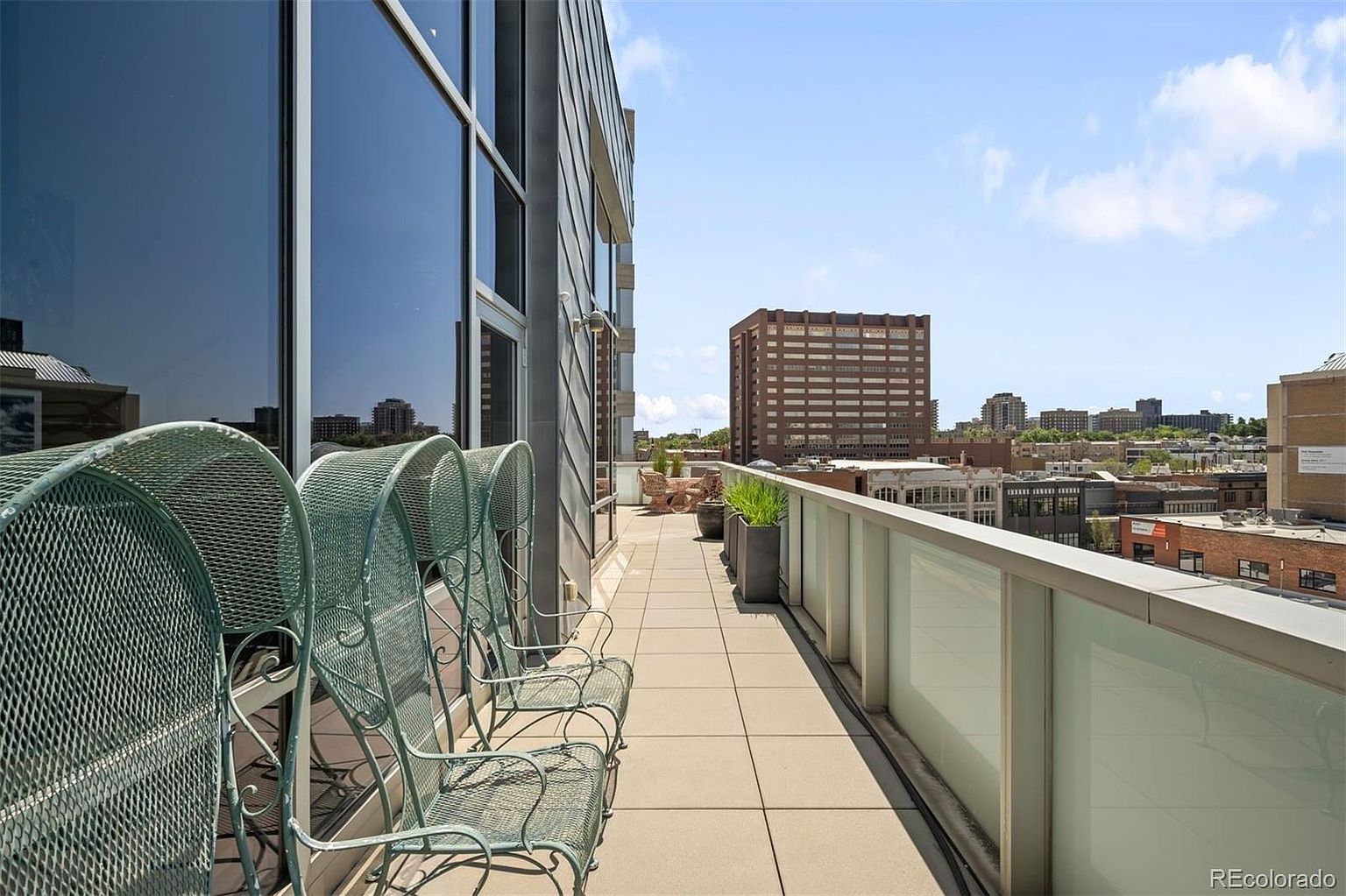 This image showcases a spacious balcony with a cityscape view. The balcony features tiled flooring, a glass railing, and several metal chairs, creating an inviting outdoor living space. The building's modern architecture is visible, with large windows reflecting the sky.