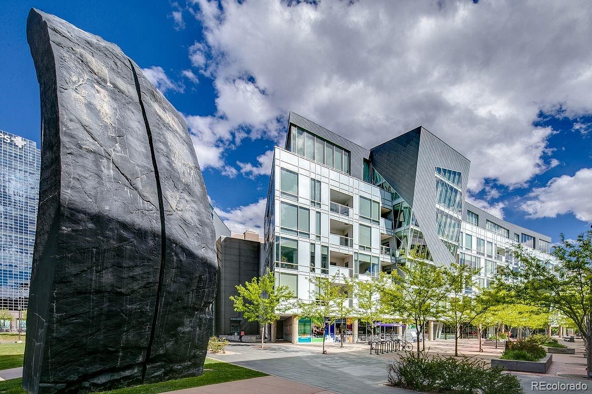 This image showcases the front exterior of a modern building with unique architectural design, featuring a mix of glass and metal elements. A large, dark rock sculpture stands prominently in the foreground, adding an artistic touch to the property's entrance. Lush greenery and a paved walkway lead to the building, creating an inviting and contemporary urban landscape.