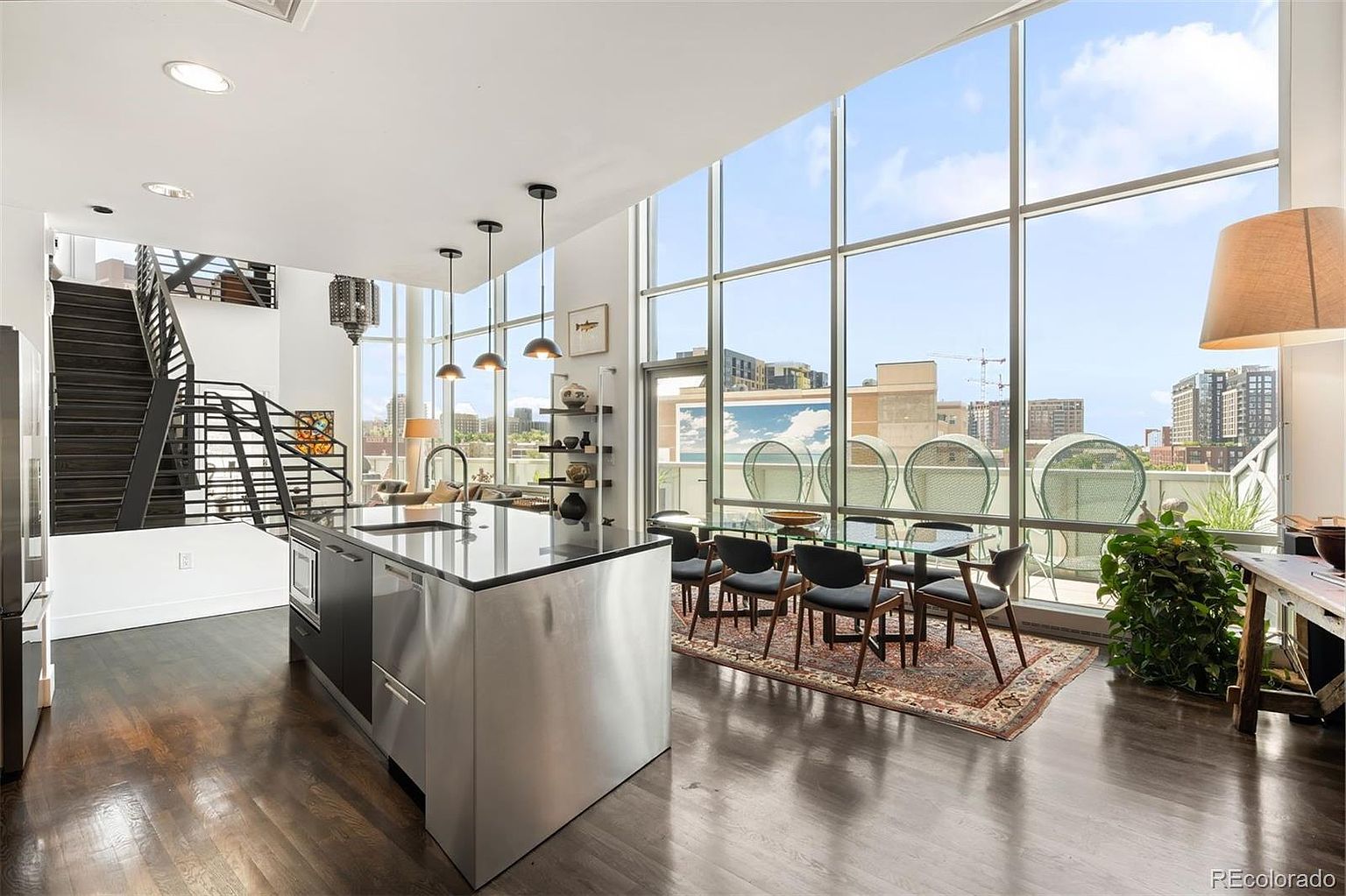 This is a bright and modern kitchen and dining area with floor-to-ceiling windows offering city views. The kitchen features a stainless steel island with a dark countertop, while the dining area includes a glass table and contemporary chairs, complemented by an area rug. A staircase is visible in the background, adding architectural interest to the open-concept space.