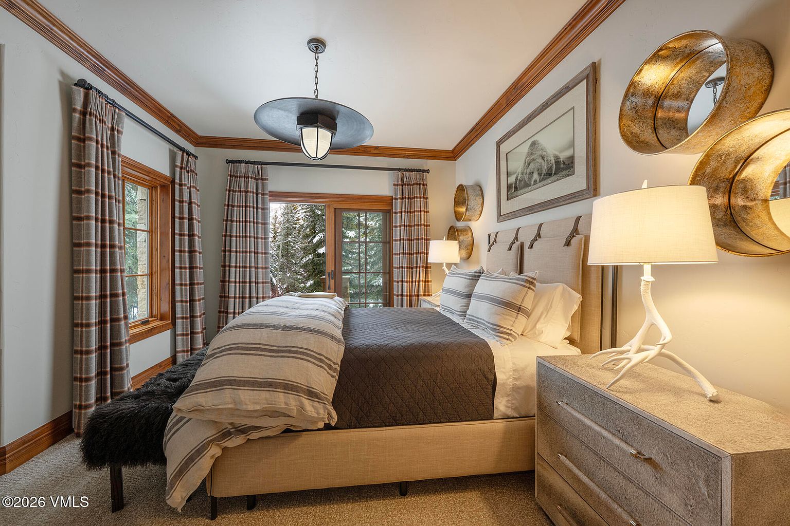 This cozy bedroom features a neutral color palette with rustic mountain-inspired decor, including plaid curtains and textured wall art. A plush bed with striped bedding serves as the focal point, flanked by a bedside table with a unique antler-style lamp. The room is illuminated by a large, industrial-style pendant light and offers a view of a snowy forest through the sliding glass doors.