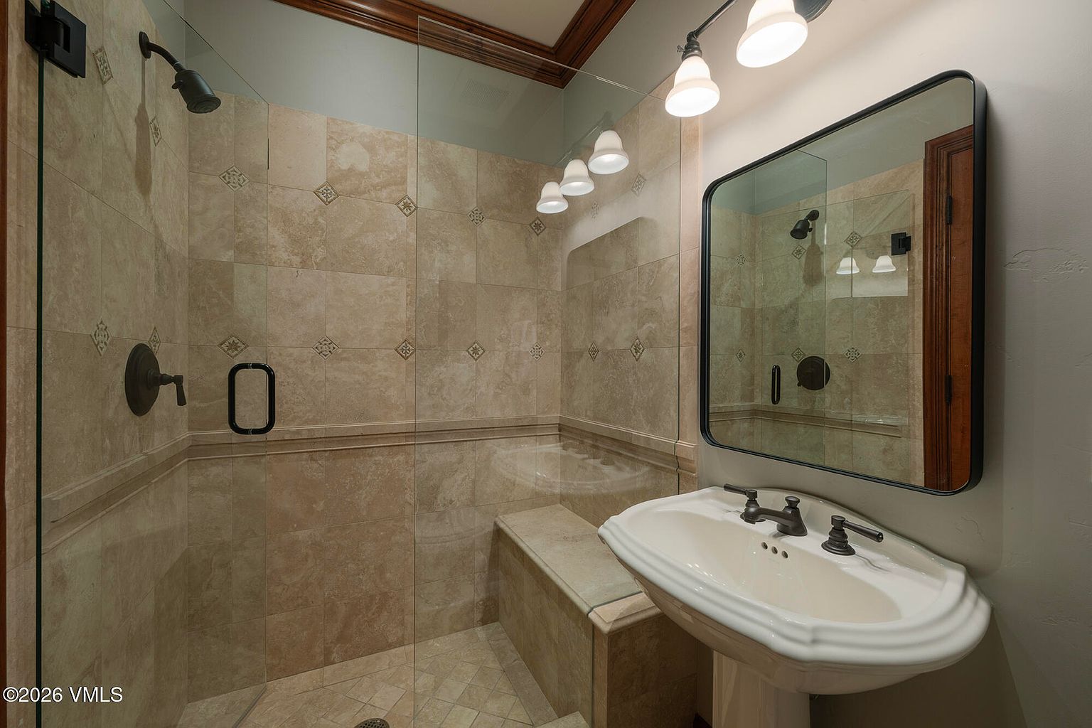 This compact bathroom features a walk-in shower enclosed by glass panels, showcasing neutral-toned stone tile work and a built-in bench. A pedestal sink with dark hardware sits adjacent to the shower, complemented by a large, dark-framed mirror and overhead vanity lighting. The space offers a clean, functional design with a cohesive color palette.