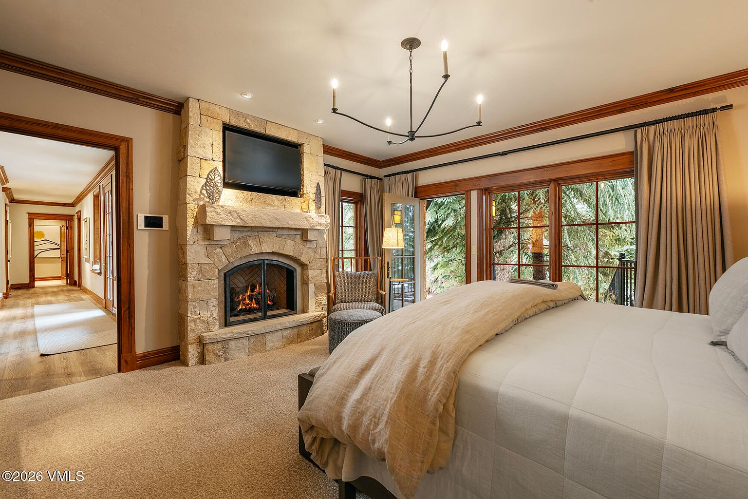 This primary bedroom features a cozy, rustic aesthetic centered around a stone-clad chimney breast with a built-in fireplace and television. A plush bed with neutral bedding sits in the foreground, while large windows and a glass door provide views of the surrounding trees and natural light. The room is accented by warm wood trim, a modern chandelier, and a hallway leading to an adjacent space.