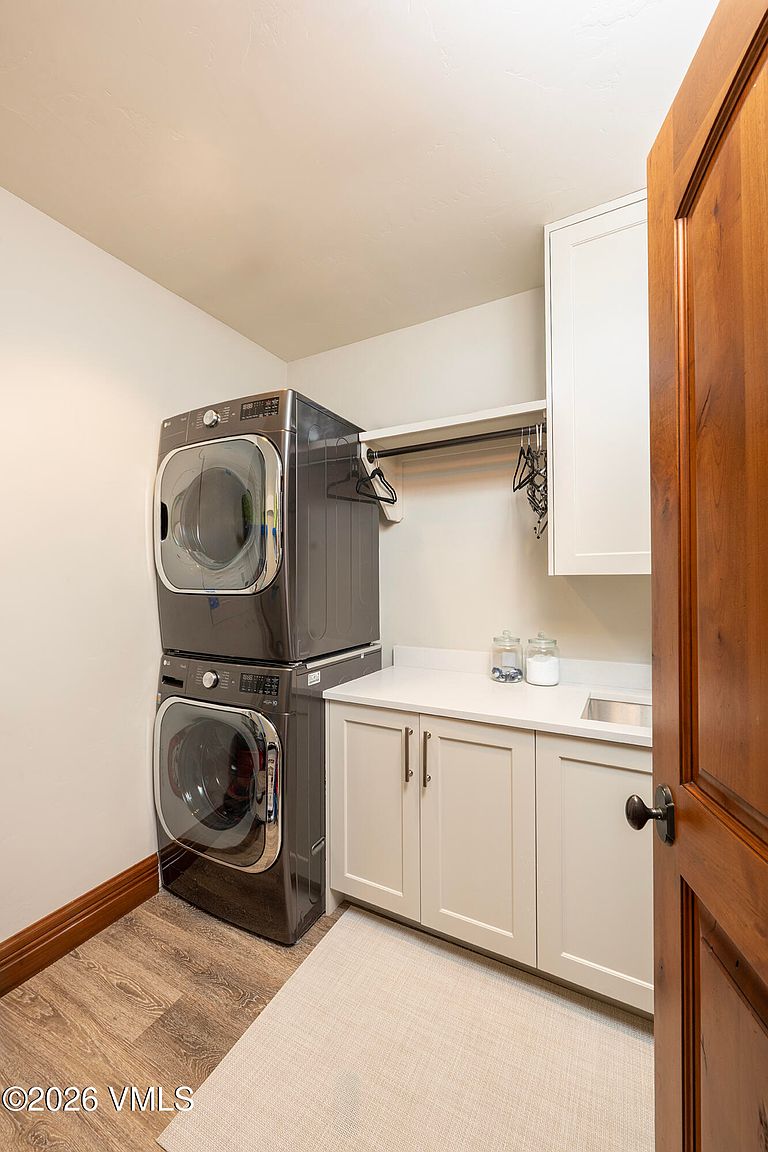 This functional laundry room features a stacked washer and dryer set in a dark metallic finish, positioned next to a white cabinet base with a light-colored countertop and an integrated sink. A hanging rod is mounted above the counter for garment storage, and the room is finished with wood-look flooring and neutral walls. The perspective is from the doorway, looking into the compact and organized utility space.