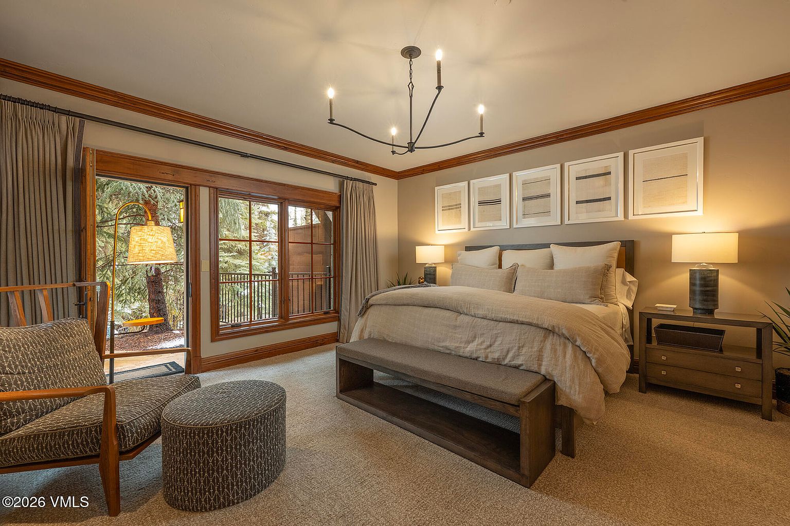 This primary bedroom features a neutral color palette with a plush bed centered against a wall adorned with five framed art pieces. Large windows with wooden trim provide a view of the surrounding trees, while a comfortable armchair and ottoman sit in the foreground. The room is illuminated by a modern chandelier and matching bedside lamps, creating a warm and inviting atmosphere.