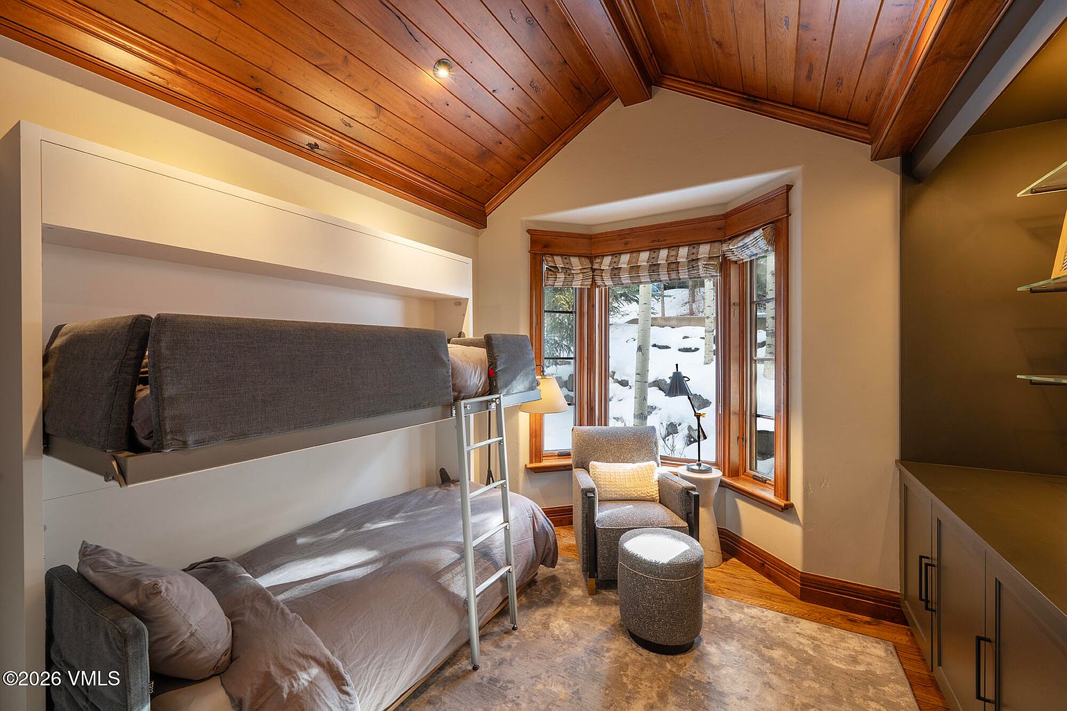 This cozy bedroom features a built-in bunk bed setup with a dark gray upholstered frame and a metal ladder. A comfortable armchair and matching ottoman are positioned near a bay window that offers a view of a snowy, wooded landscape. The room is accented by a warm wood-paneled vaulted ceiling and a neutral-toned area rug, creating a rustic and inviting atmosphere.