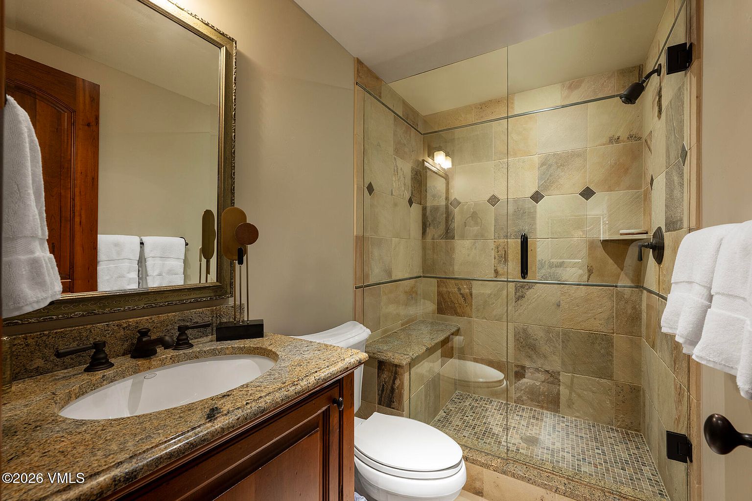 This compact bathroom features a vanity with a stone countertop and an integrated sink, paired with a large framed mirror. To the right, a glass-enclosed shower stall is finished with textured stone wall tiles and a mosaic tile floor, including a built-in stone bench. The space is completed by a white toilet, creating a functional and cohesive design.