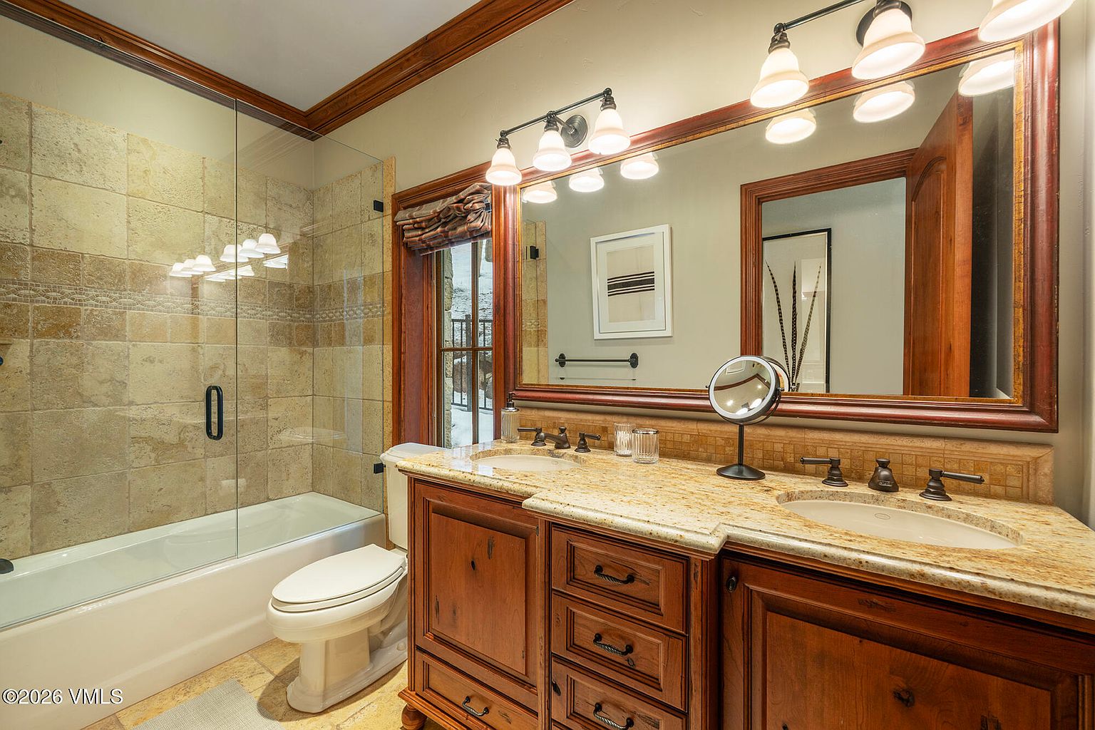 This primary bathroom features a dual-sink vanity with a light-toned stone countertop and warm-toned wood cabinetry. A large, framed mirror spans the vanity area, complemented by a glass-enclosed shower with neutral-toned wall tiles. The space is illuminated by a multi-light fixture above the mirror and includes a toilet positioned next to the shower.