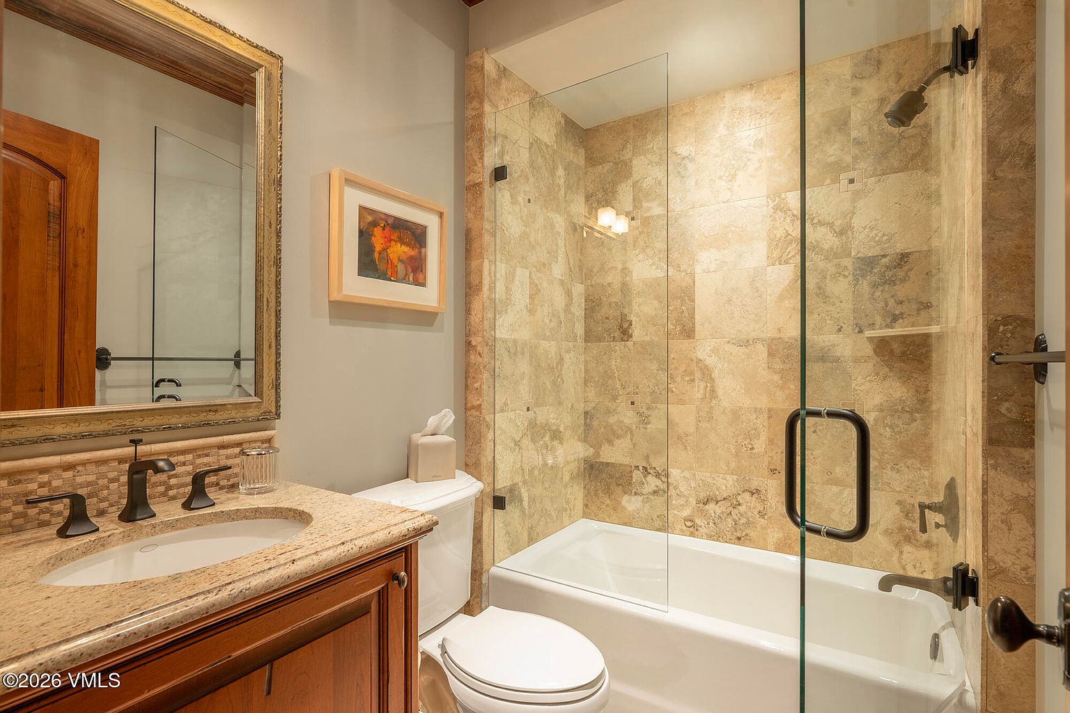 This compact bathroom features a vanity with a stone countertop, a framed mirror, and a combination tub and shower enclosure. The shower area is finished with neutral-toned square tiles and enclosed by a glass panel with a dark metal handle. The space is painted in a soft grey tone, creating a clean and functional aesthetic.