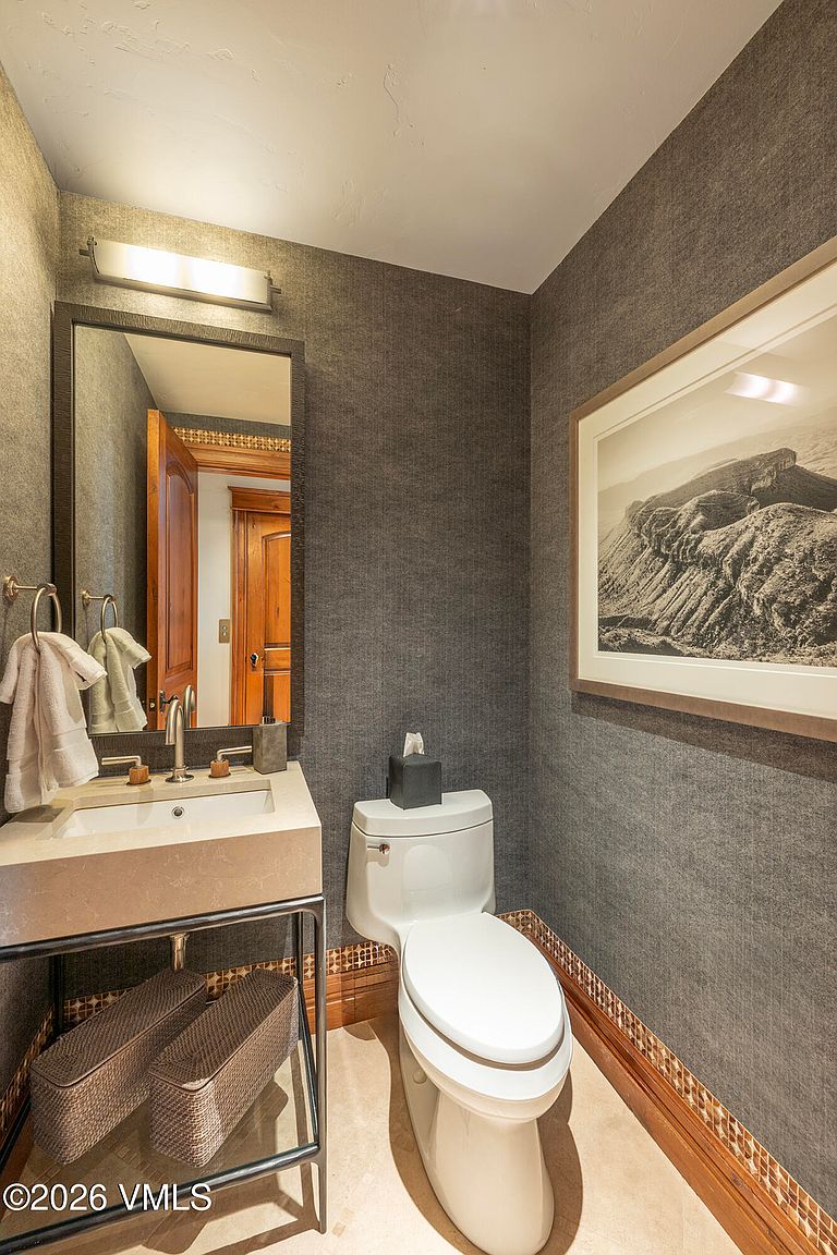 This elegant powder room features dark, textured wallpaper and a sleek, wall-mounted stone vanity with a metal frame base. A framed landscape photograph hangs on the wall, while a decorative tile border accents the base of the walls near the floor. The space is completed by a modern toilet and a rectangular mirror illuminated by a minimalist light fixture.
