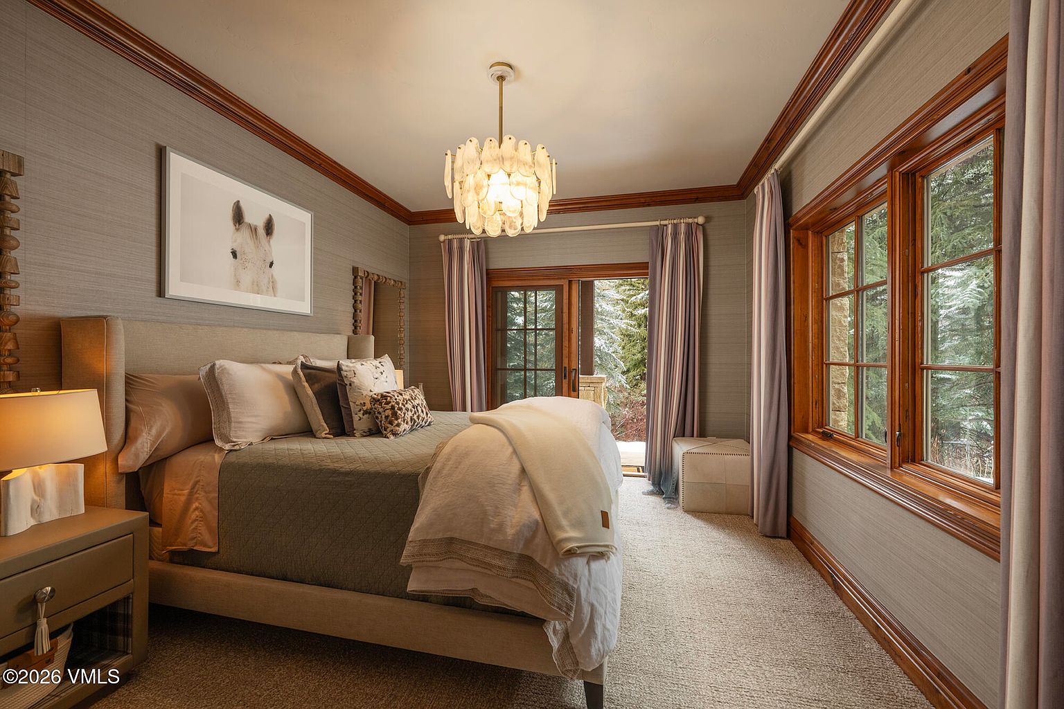 This elegant primary bedroom features a neutral color palette with textured wall coverings and rich wood trim around the windows and crown molding. A plush bed with layered linens serves as the focal point, positioned beneath a decorative chandelier, while large windows and a glass door provide views of a snowy outdoor landscape. The room is completed by a bedside table with a lamp and a small ottoman, creating a cozy and sophisticated retreat.