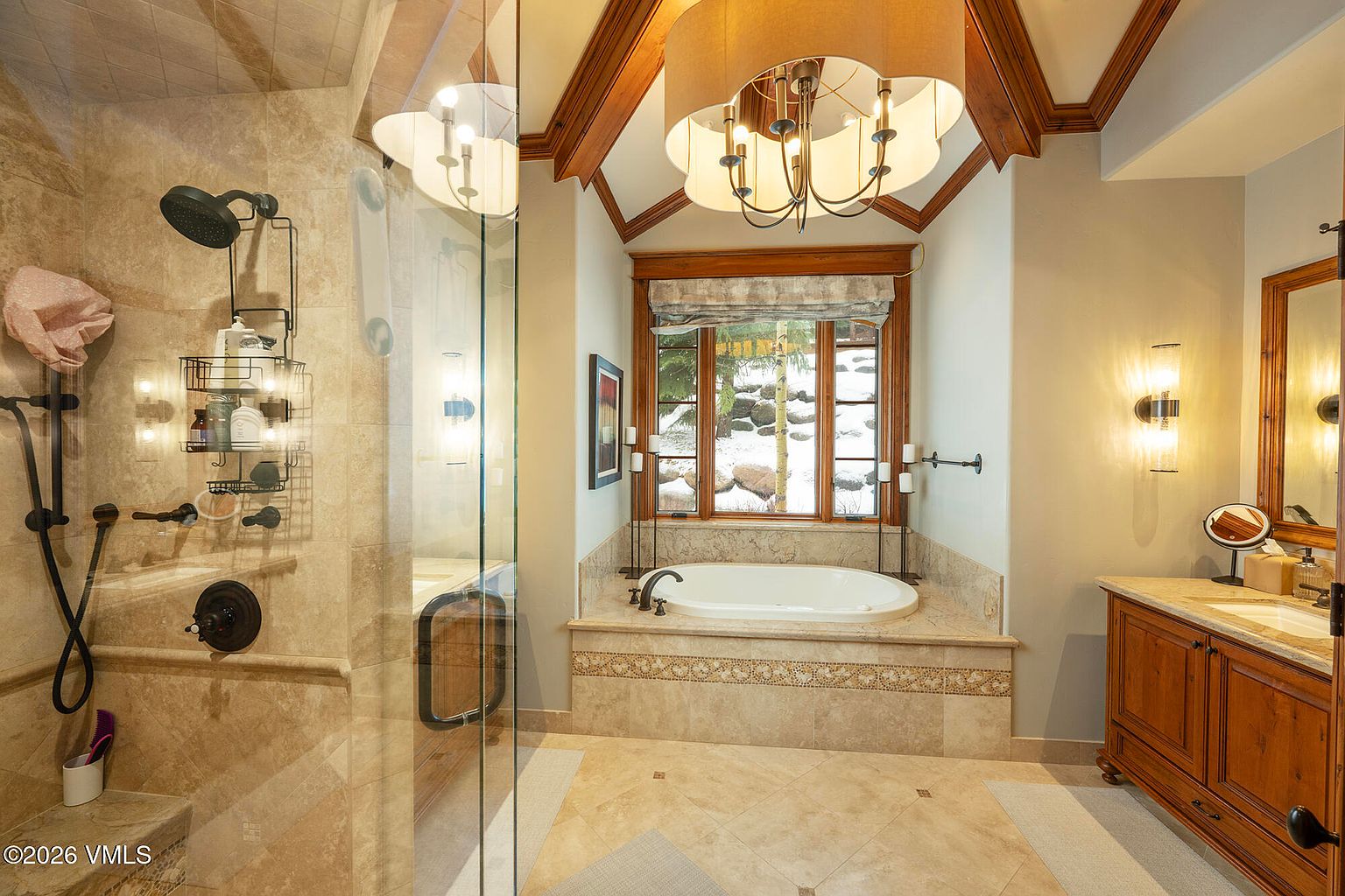 This luxurious primary bathroom features a large soaking tub nestled in a tiled alcove beneath a window with a snowy outdoor view. To the left, a glass-enclosed walk-in shower is equipped with multiple showerheads, while a wooden vanity with a sink and mirror occupies the right side. The room is accented by a decorative chandelier hanging from a vaulted ceiling with wood trim, creating an elegant and warm atmosphere.
