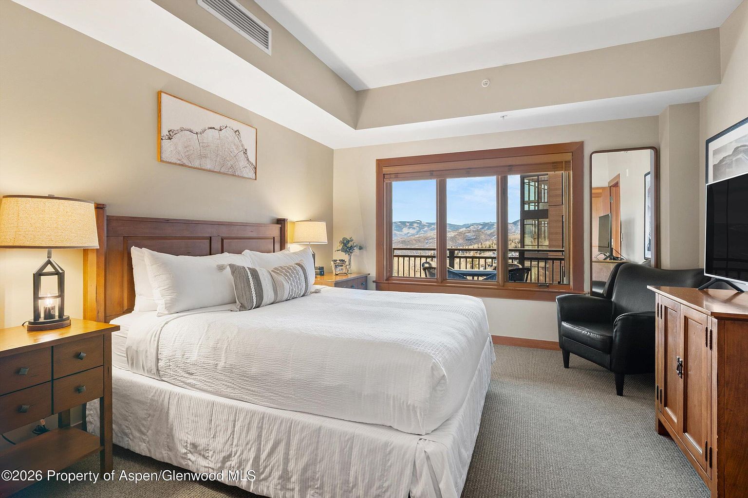 This is a primary bedroom featuring a king-size bed with white linens and decorative pillows. The room has a neutral color palette with a wooden headboard and matching nightstands. A large window offers a view of the mountains, and there is a comfortable armchair and a wooden dresser with a television.