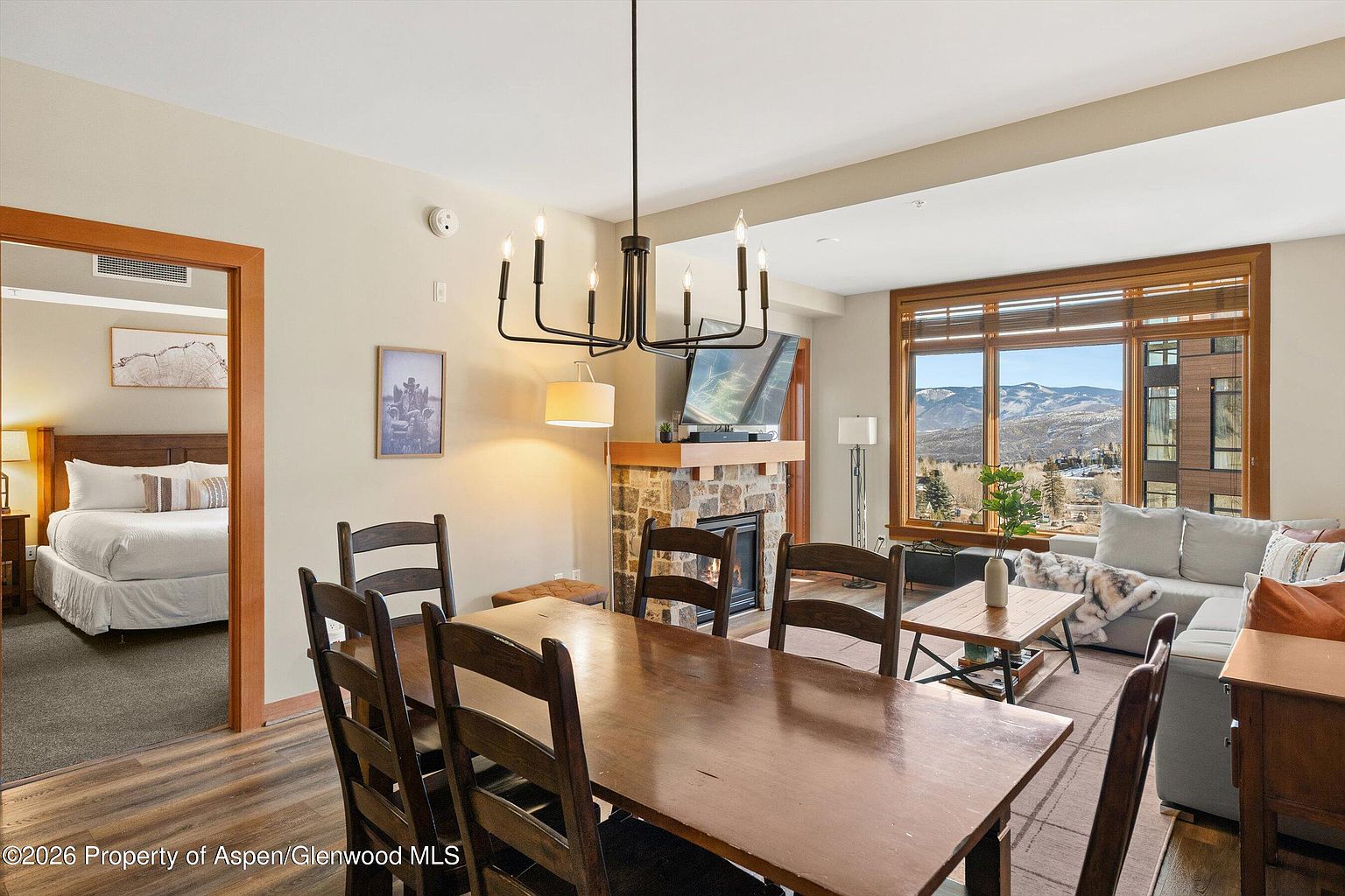 This interior shot showcases a dining room with a large wooden table and chairs, illuminated by a modern chandelier. A stone fireplace with a television above it adds a cozy focal point, while a large window provides a scenic mountain view. The room seamlessly connects to a living area, creating an open and inviting space.