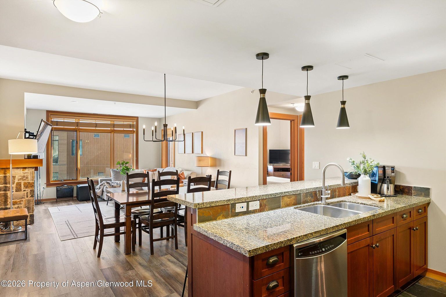This interior shot showcases a well-appointed kitchen that seamlessly blends into the dining and living areas. The kitchen features granite countertops, wooden cabinetry, and stainless steel appliances, while the dining area includes a wooden table with chairs and a modern chandelier. The living area is visible in the background, featuring a stone fireplace and large windows, creating an open and inviting living space.