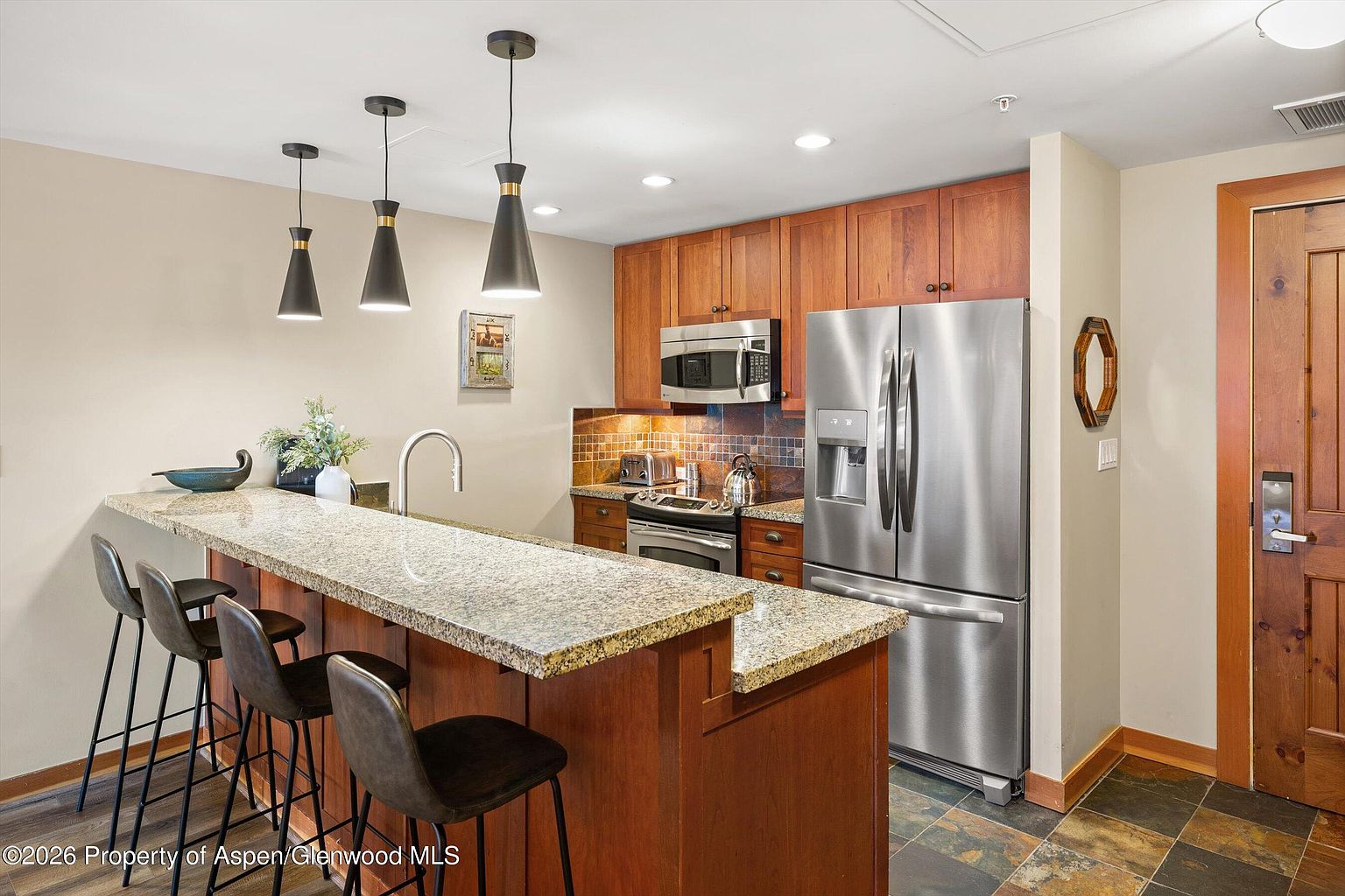 This is a well-lit kitchen featuring wooden cabinetry, stainless steel appliances, and a granite countertop island with bar seating. The kitchen has a warm and inviting feel, with modern pendant lighting and a tiled backsplash adding visual interest. The flooring appears to be slate tile.