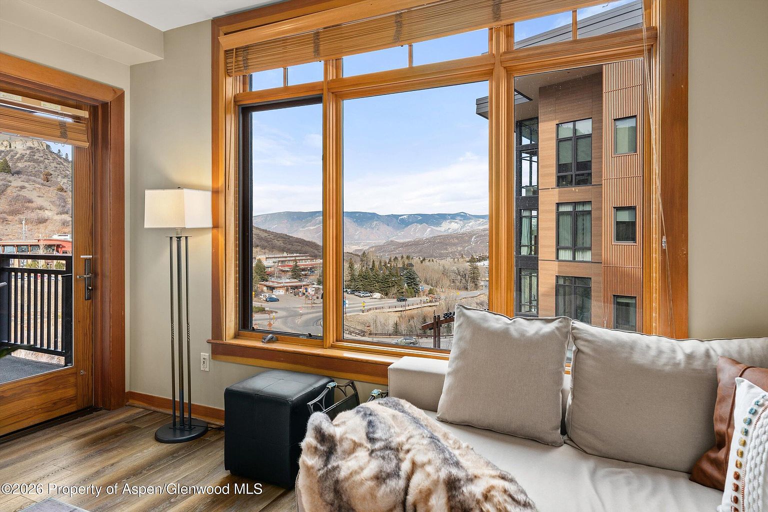 This interior shot showcases a cozy living room with a large window offering a scenic view of mountains and a nearby town. The room features a comfortable sofa with plush pillows and a throw blanket, complemented by a floor lamp and a dark ottoman. The hardwood flooring and wooden window frames add warmth to the space, creating an inviting atmosphere.