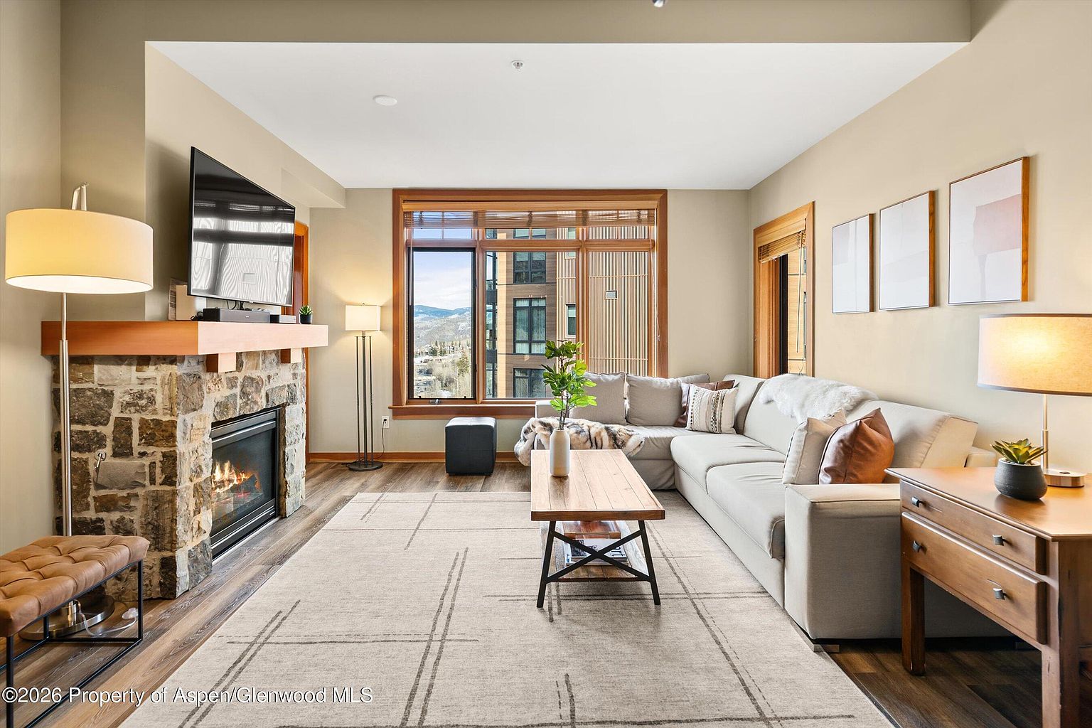 This is an interior shot of a living room featuring a stone fireplace with a mounted television above it. A large sectional sofa is positioned near a window with wooden trim, and a wooden coffee table sits on a patterned rug. The room is decorated in neutral tones, creating a warm and inviting atmosphere.