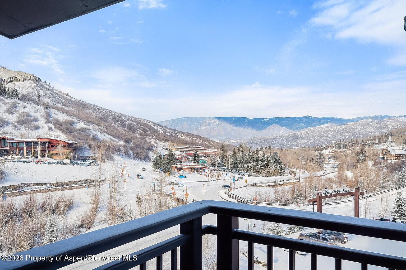 This image showcases a scenic view from a balcony, overlooking a snow-covered landscape with mountains in the background. The balcony features a dark railing, providing a clear view of the surrounding area, including buildings, roads, and trees dusted with snow. The perspective is from the balcony, offering a sense of immersion in the winter scenery.