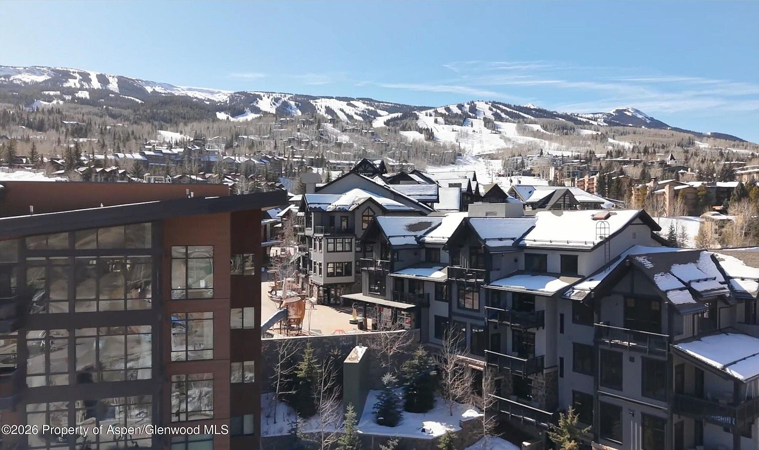 This aerial view showcases a luxurious mountain resort community with snow-covered rooftops and stunning mountain vistas in the background. The architecture features a mix of modern and traditional styles, with multi-story buildings and private balconies. The scene conveys a sense of upscale living and access to world-class skiing.