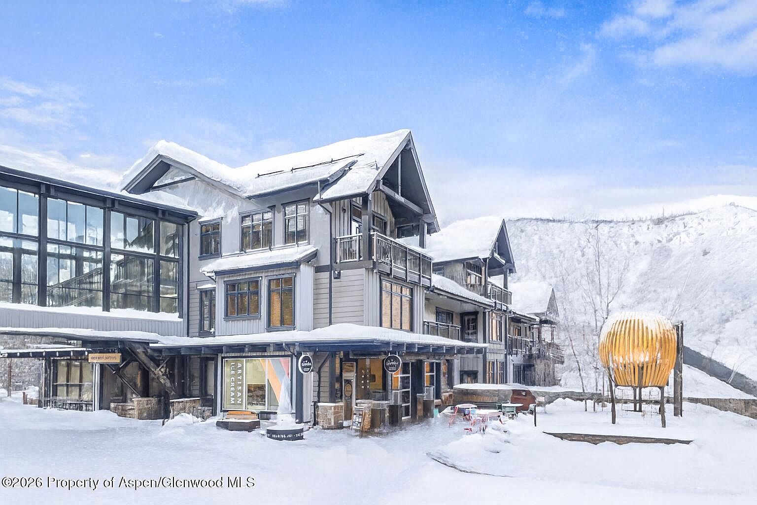 This image showcases the front exterior of a multi-story building complex in a snowy, mountainous setting. The architecture features a blend of modern and rustic elements, with gray siding, dark trim, and snow-covered roofs. The building includes commercial spaces on the ground floor, indicated by storefronts with signage, and residential units above, some with balconies. A unique, spherical art installation stands in the foreground, adding a touch of contemporary design to the winter landscape.