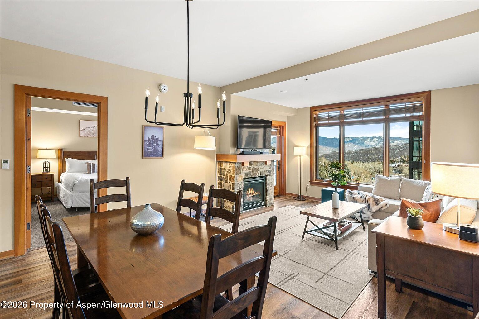 This is an interior shot showcasing a combined living and dining area. The living room features a stone fireplace with a mounted TV, a comfortable sofa facing a large window with mountain views, and a wooden side table. The dining area includes a wooden table with chairs and a modern chandelier, with a glimpse into a bedroom through an open doorway. The overall impression is a cozy and inviting space with a blend of rustic and contemporary elements.