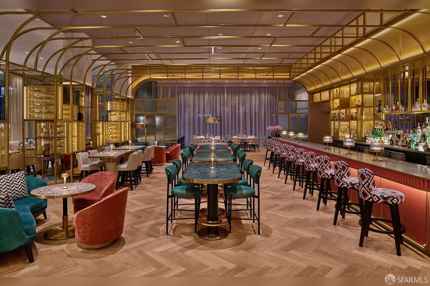 This sophisticated lounge or bar area features a high-end, modern aesthetic with a prominent gold-toned metal framework overhead. The space is furnished with a long bar, high-top tables with dark stone surfaces, and plush, colorful velvet seating, all set against a backdrop of herringbone-patterned wood flooring. The lighting is warm and ambient, creating an inviting atmosphere suitable for a luxury residential amenity space.