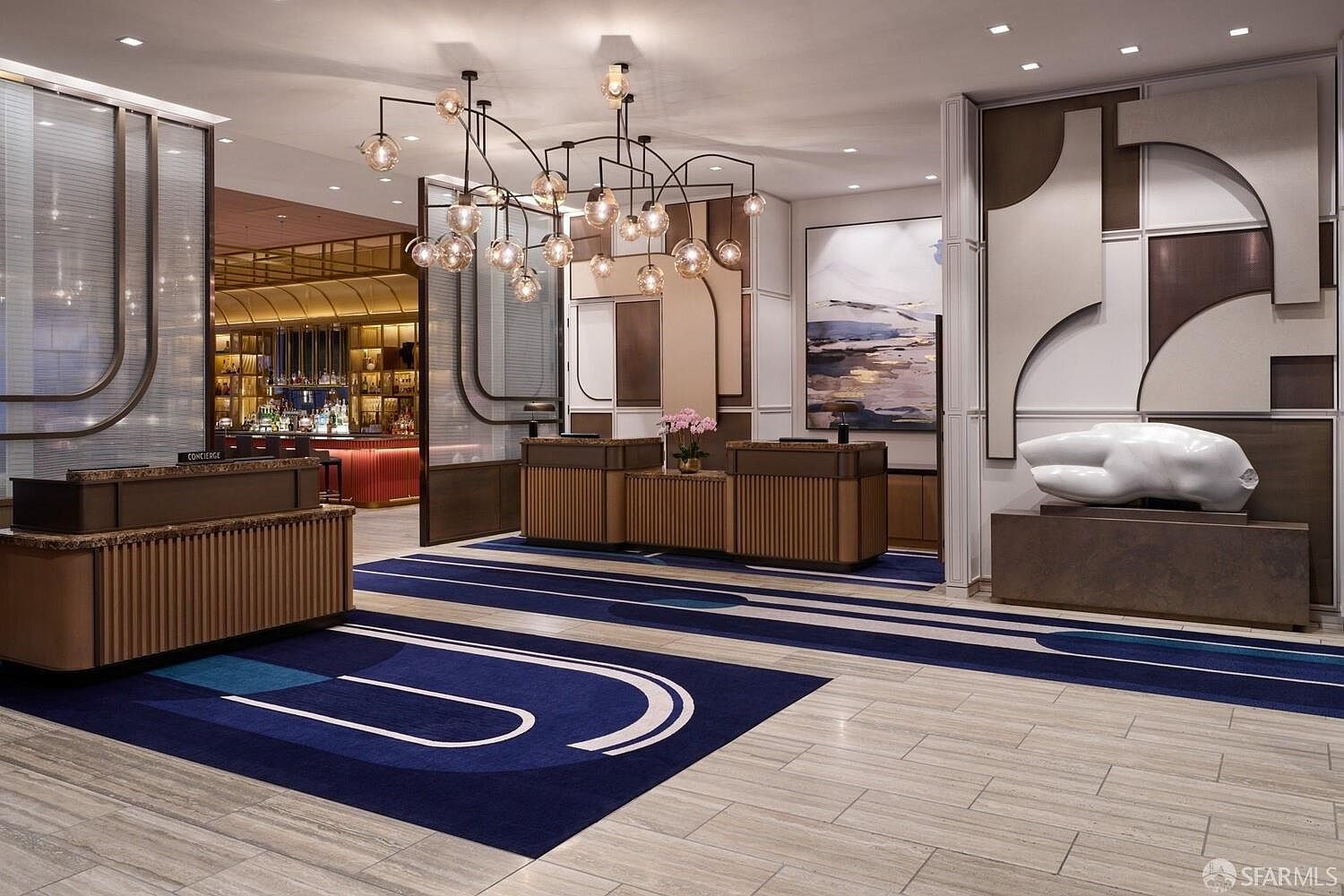 This sophisticated hotel lobby features a grand reception area with multiple service desks and a striking, modern chandelier as the focal point. The space is defined by a bold, geometric blue and white patterned carpet, warm wood-toned accents, and a large abstract sculpture on a pedestal. In the background, a glimpse of a well-stocked bar area adds to the upscale, professional atmosphere of this commercial space.